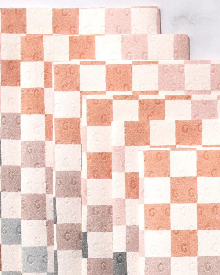 Five overlapping GEOMETRY Autumn Checkers towels show a checkered beige, peach, and gray pattern on white. Each square is embossed with a “G,” combining eco-friendly style for modern sustainable kitchens.