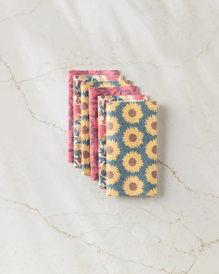 Four folded "Autumn Florals" reusable paper towels by Rebecca Bobko lay on a marble surface: two with a blue and yellow sunflower print, and two with a pink bird and flower pattern.