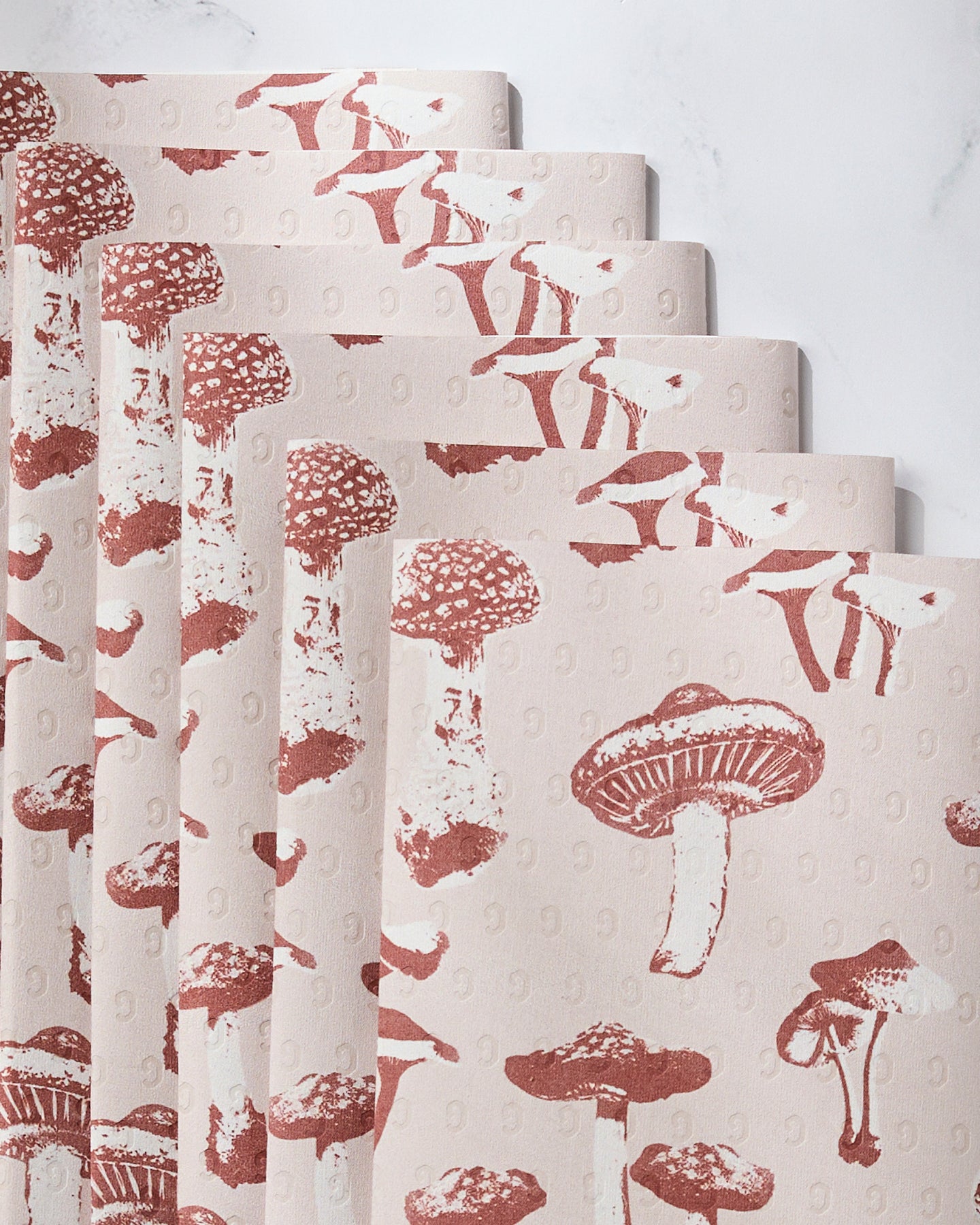 A fanned stack of Geometry’s Autumn Mushrooms Not Paper Towels, featuring a repeating red mushroom pattern, displayed on light marble—an eco-friendly and stylish addition to any kitchen.
