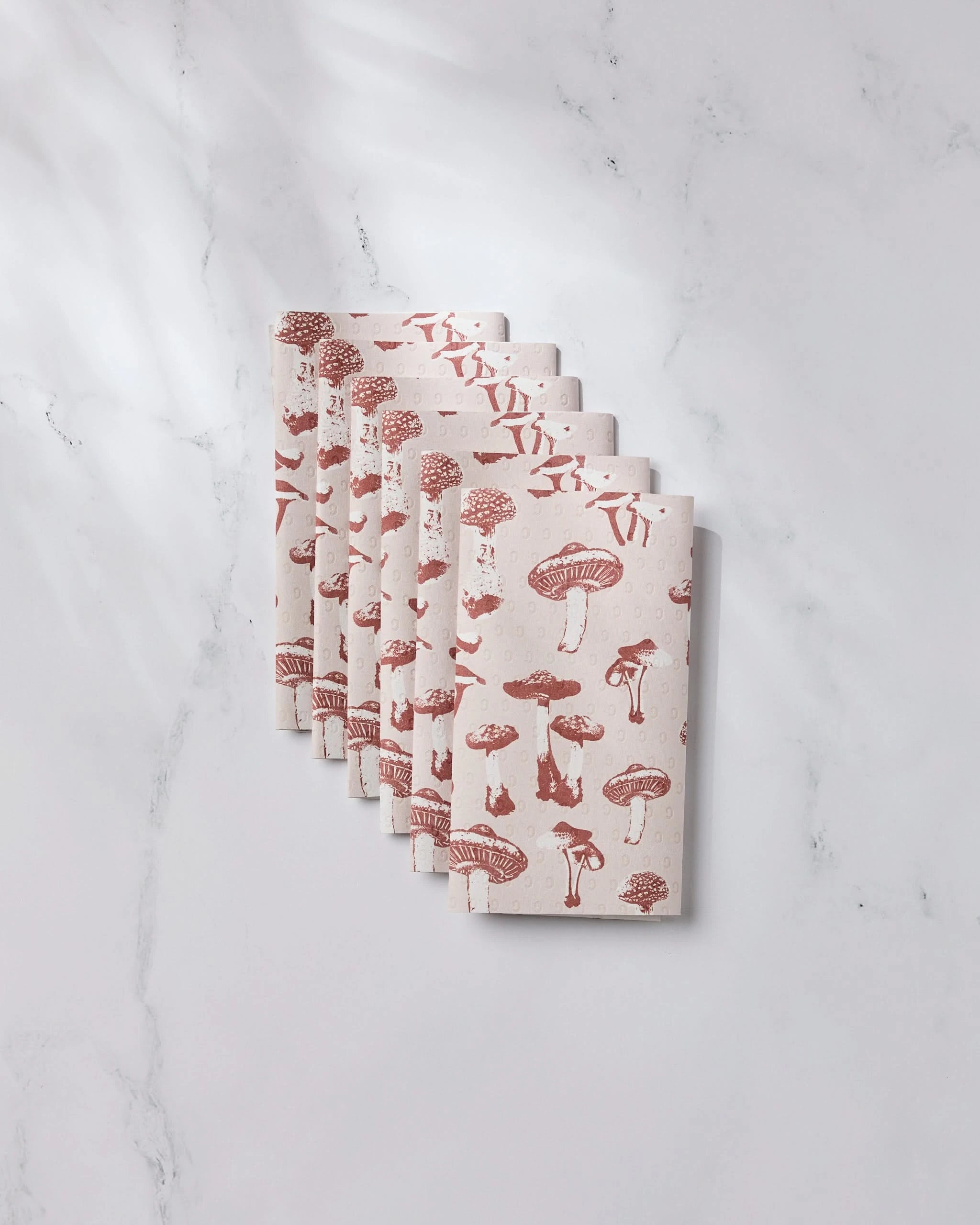 Five folded Autumn Mushrooms napkins by Geometry, featuring a red mushroom print on beige, are neatly stacked on white marble. These reusable towels add an eco-friendly touch, with soft shadows completing the scene.