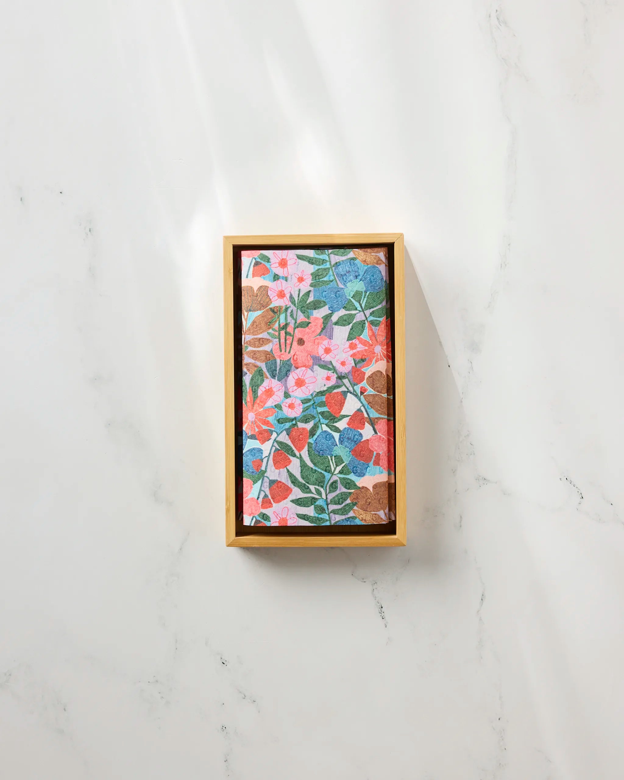 The Bloom Entangled puzzle by Lisa Nelson, featuring pink, red, and blue flowers with green leaves, is framed in a wooden box on a white marble surface beside eco-friendly paper towels.