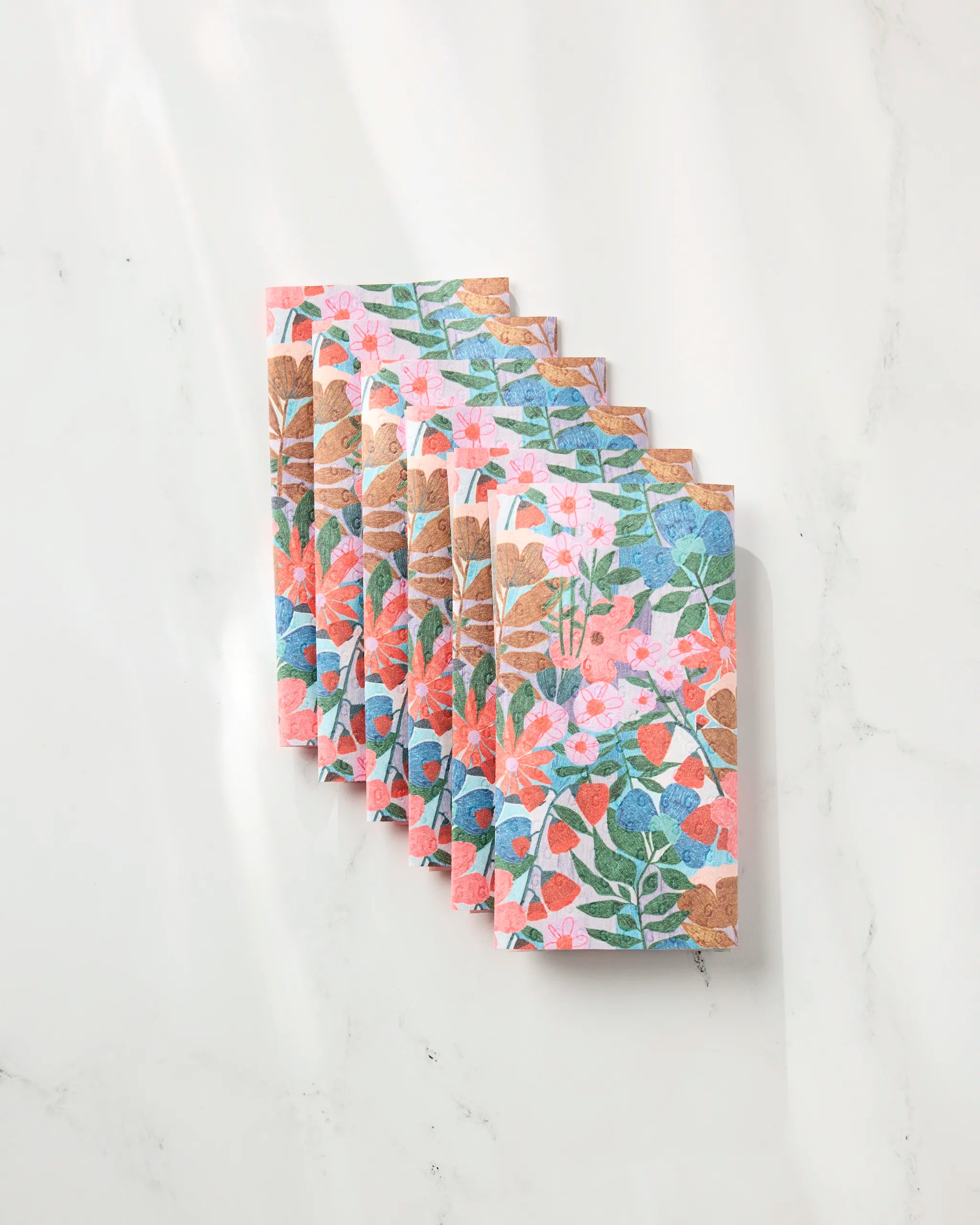 Lisa Nelson's Bloom Entangled eco-friendly paper towels, featuring vibrant pink, blue, orange, and green floral patterns, are neatly stacked on a white marble surface in gentle sunlight.