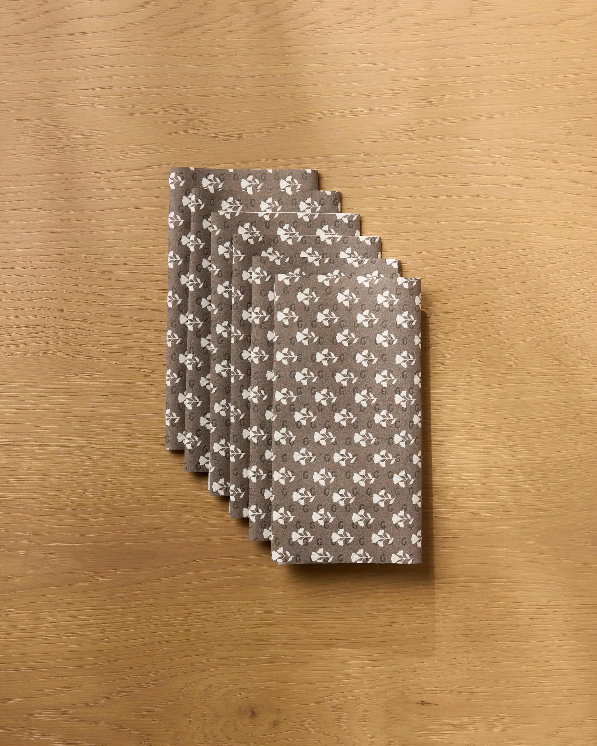 A stack of Bloom for Mom gray reusable paper towels from Geometry, featuring a white floral pattern, is arranged on a light wooden surface.