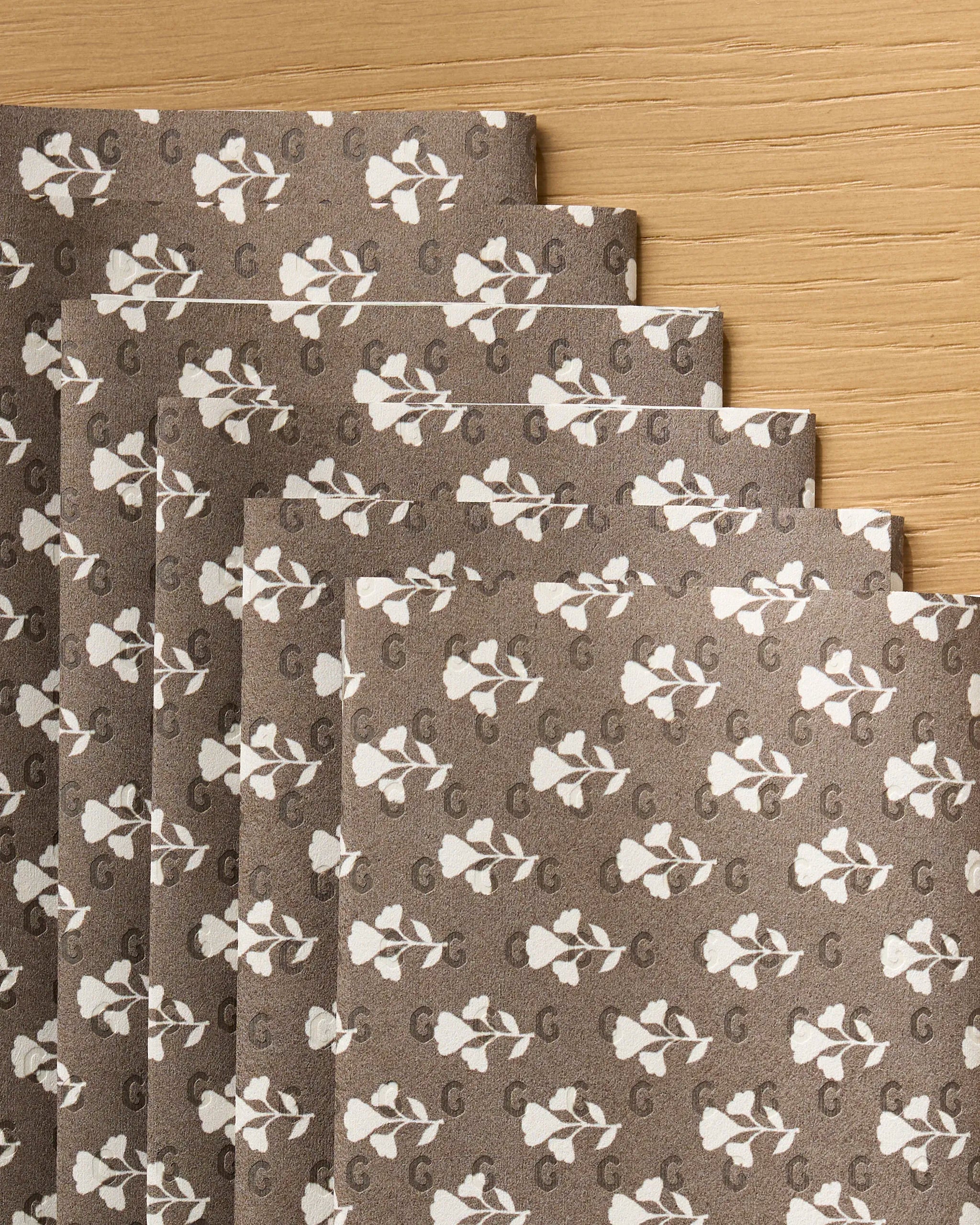Six sheets of Geometry's "Bloom for Mom" brown eco-friendly kitchen towels, featuring a white floral pattern and the letter G, are fanned out on a light wood surface.