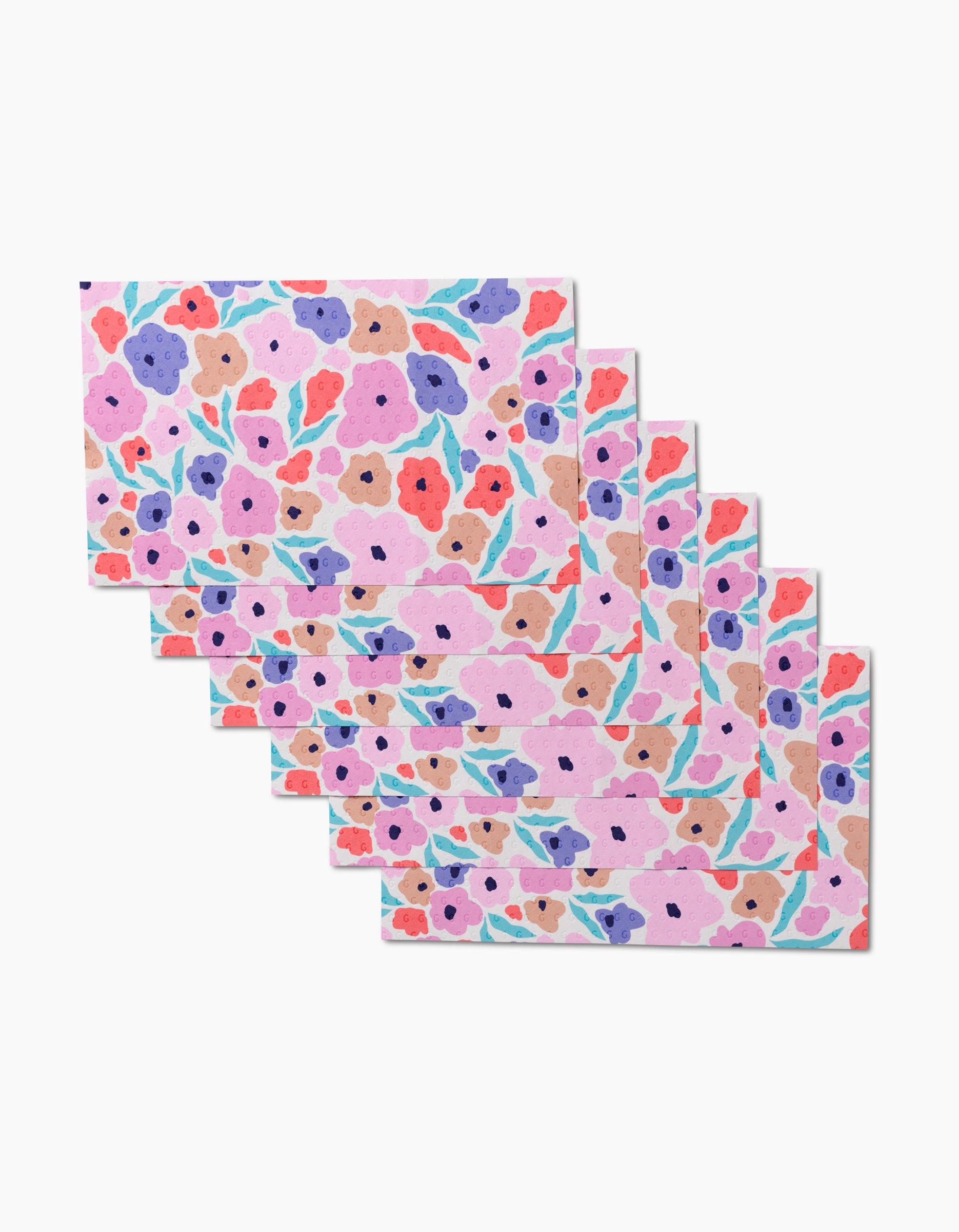 Seven “Blossoming Love” rectangular placemats by GEOMETRY, featuring a colorful floral print on light pink, made from eco-friendly post-consumer recycled materials, displayed in a staggered stack.