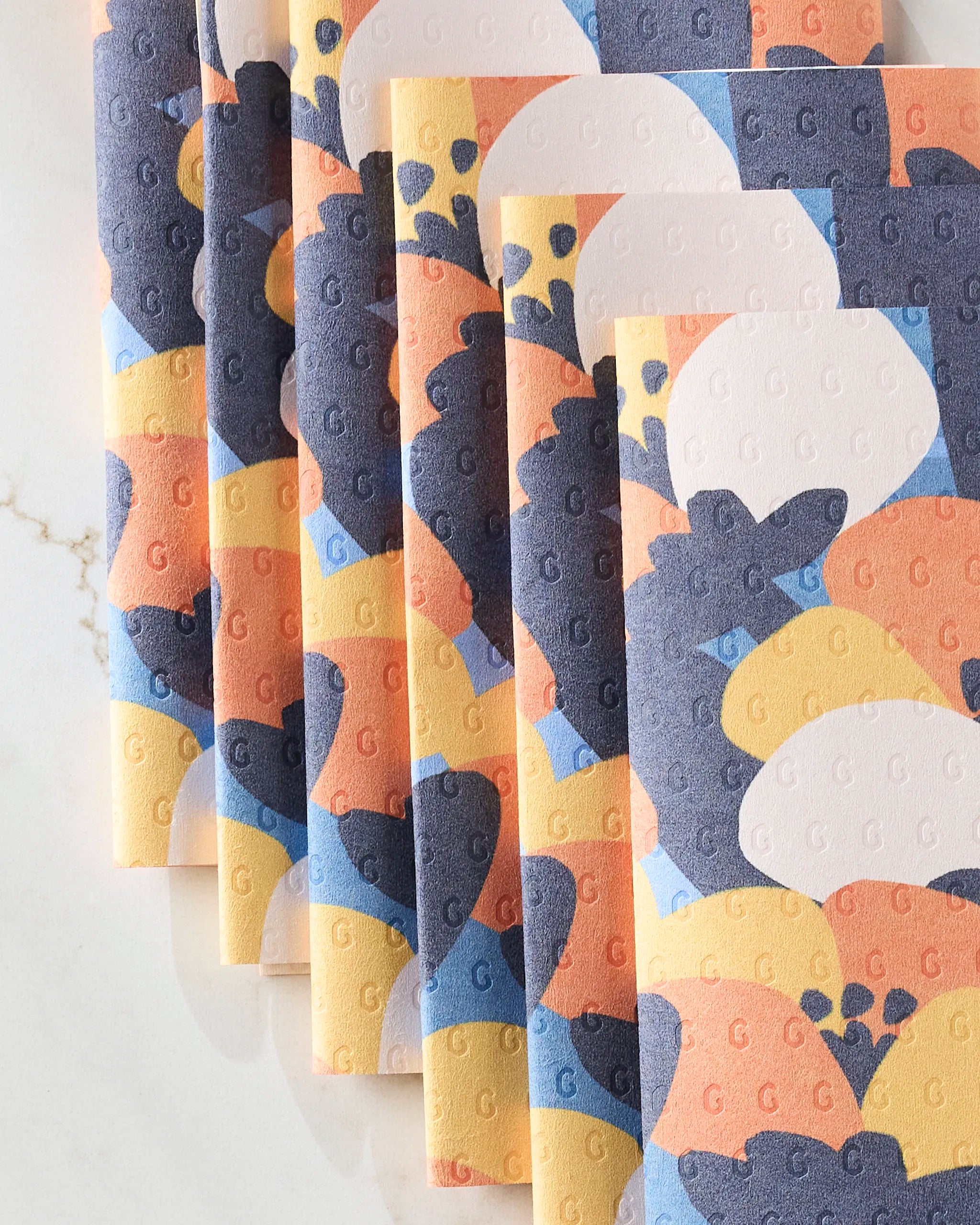 Six eco-friendly Bold Petal Bloom kitchen towels by Claire Iglesias, featuring abstract blue, orange, yellow, and white shapes, are displayed in an overlapping arrangement on a marble surface.