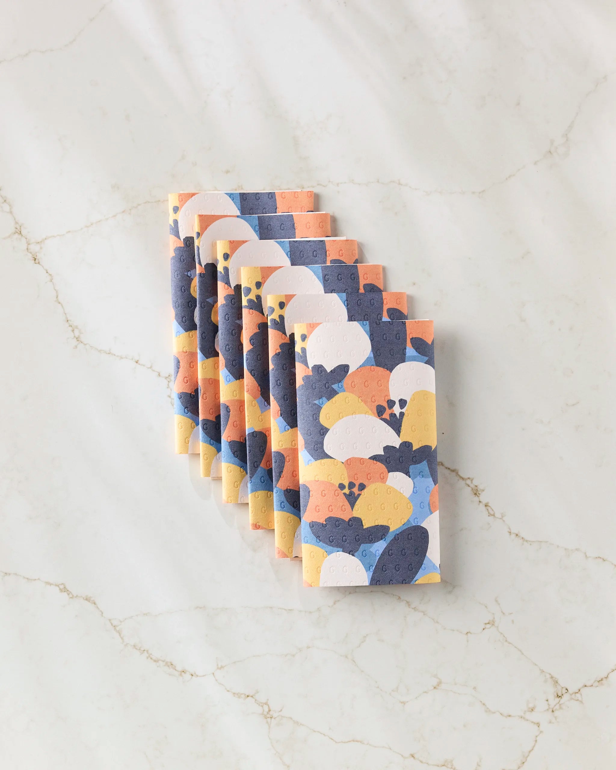 Six Bold Petal Bloom reusable paper towels by Claire Iglesias, featuring vibrant overlapping semi-circles in orange, yellow, white, and blue, are neatly stacked on a light marble surface.