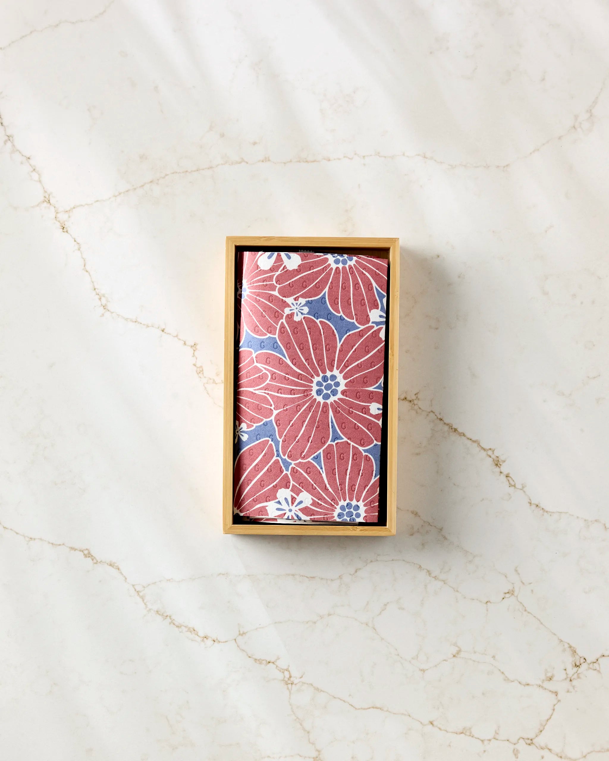 A rectangular box of Ashley Celena's Botanical Blush eco-friendly kitchen towel, featuring a red and white floral pattern on blue, sits on light marble—a reusable paper towel alternative.
