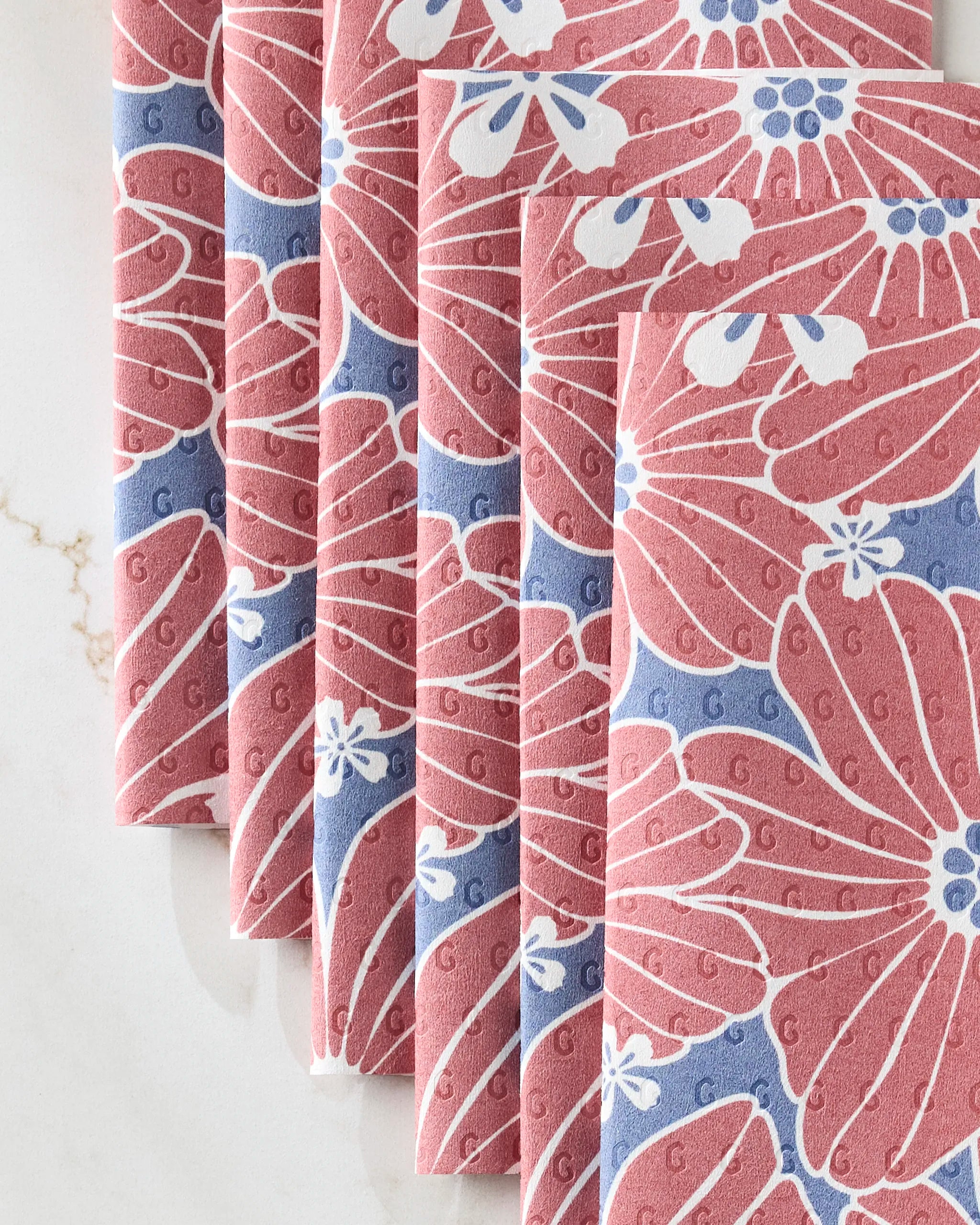 Five folded Ashley Celena "Botanical Blush" eco-friendly kitchen towels, featuring a pink and white floral pattern on blue, are neatly arranged on a light marble surface.