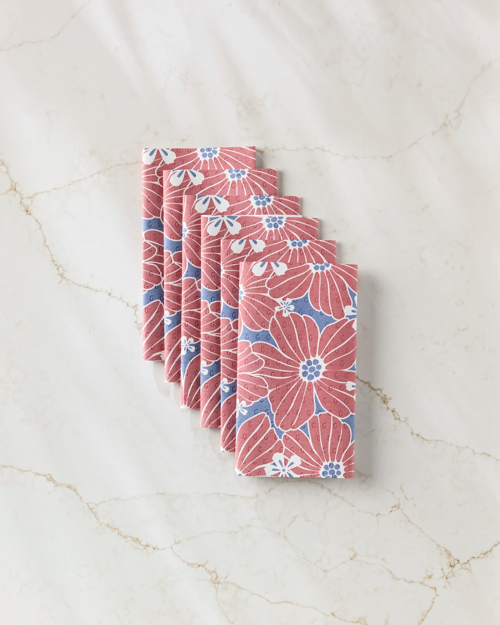 Six Botanical Blush napkins by Ashley Celena, featuring a red, blue, and white floral pattern as an eco-friendly paper towel alternative, are neatly arranged on a subtly veined light marble surface.