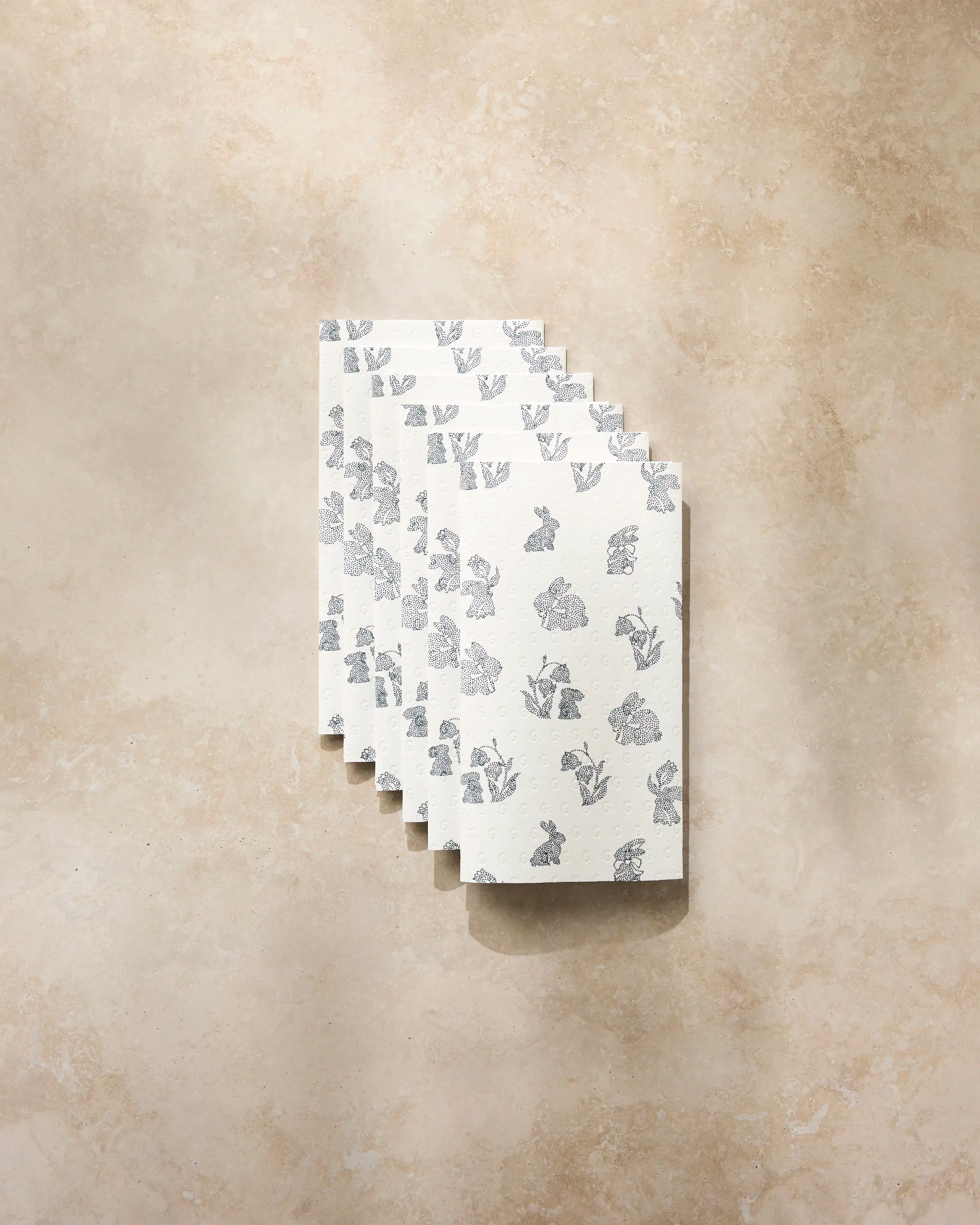 Six folded Bunny Dots napkins by Geometry, featuring a gray floral pattern, are eco-friendly paper towels arranged in a staggered row on a light beige marbled surface.