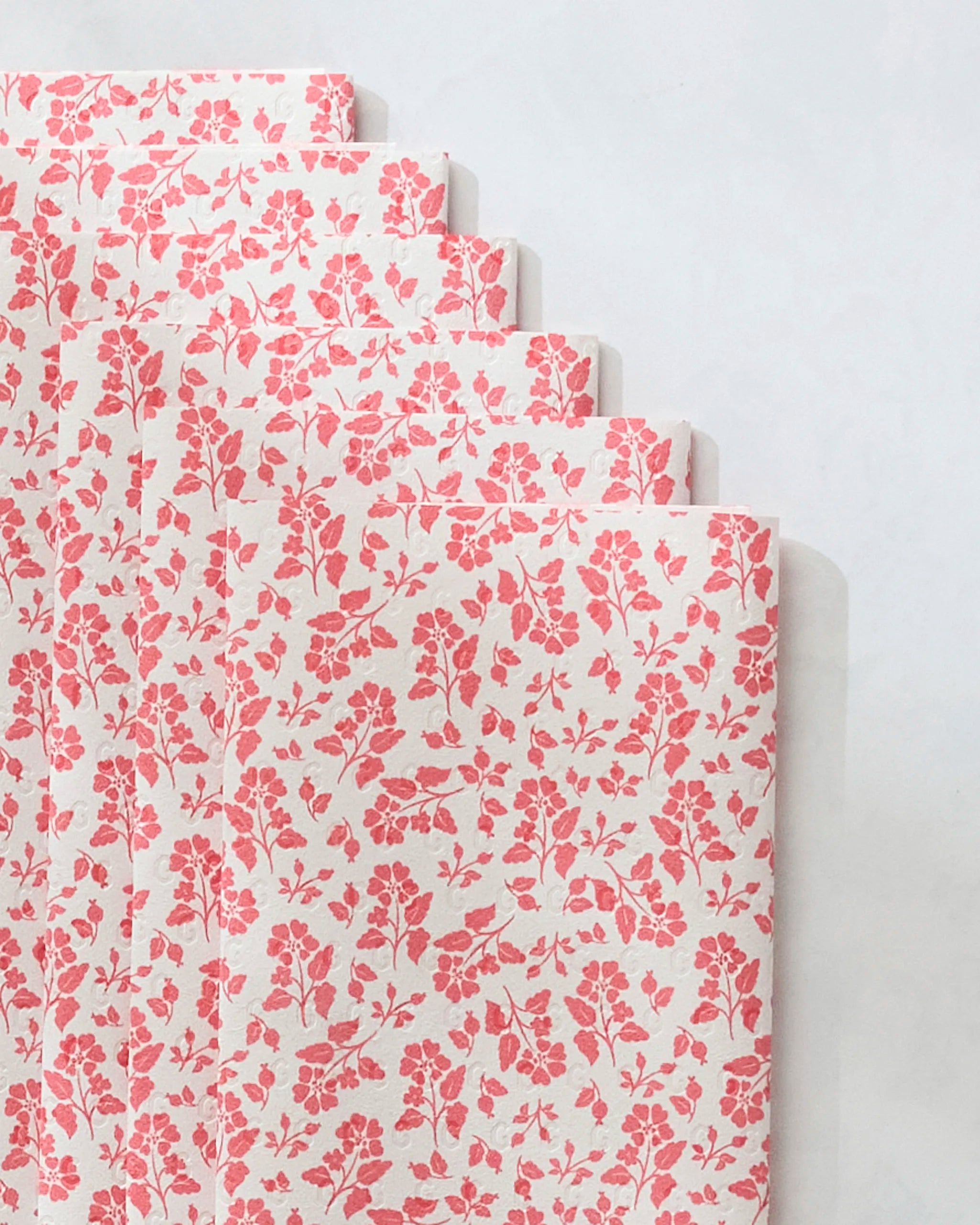 Five sheets of Danelys Sidron’s Carmine Garden eco-friendly paper towels with a pink floral pattern are stacked in a staggered arrangement against a light grey background.