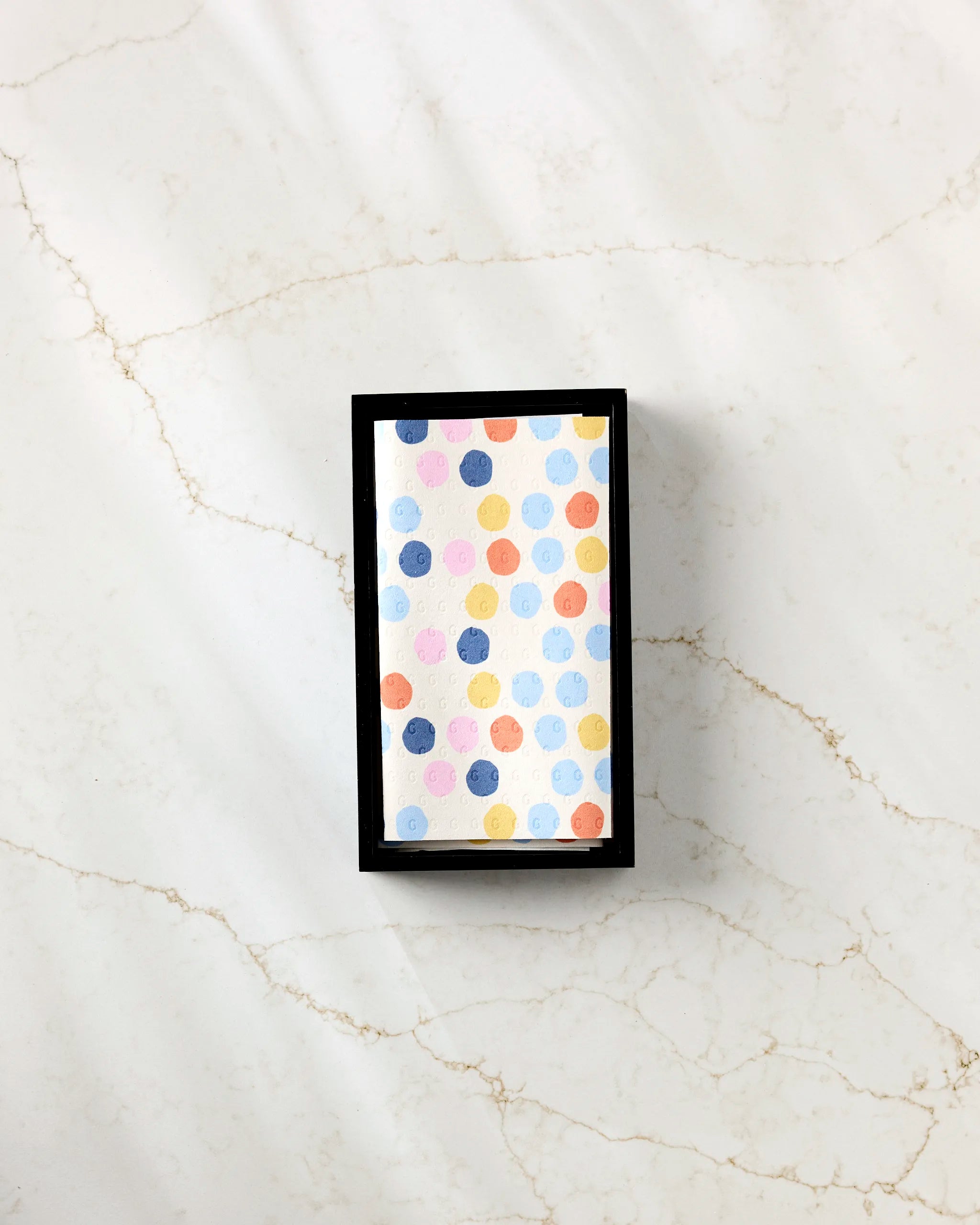 A rectangular card featuring colorful blue, orange, yellow, and red polka dots from Lizzie Clark's "Carnival" sits in a black box on white marble—evoking the playful style of eco-friendly kitchen towels.