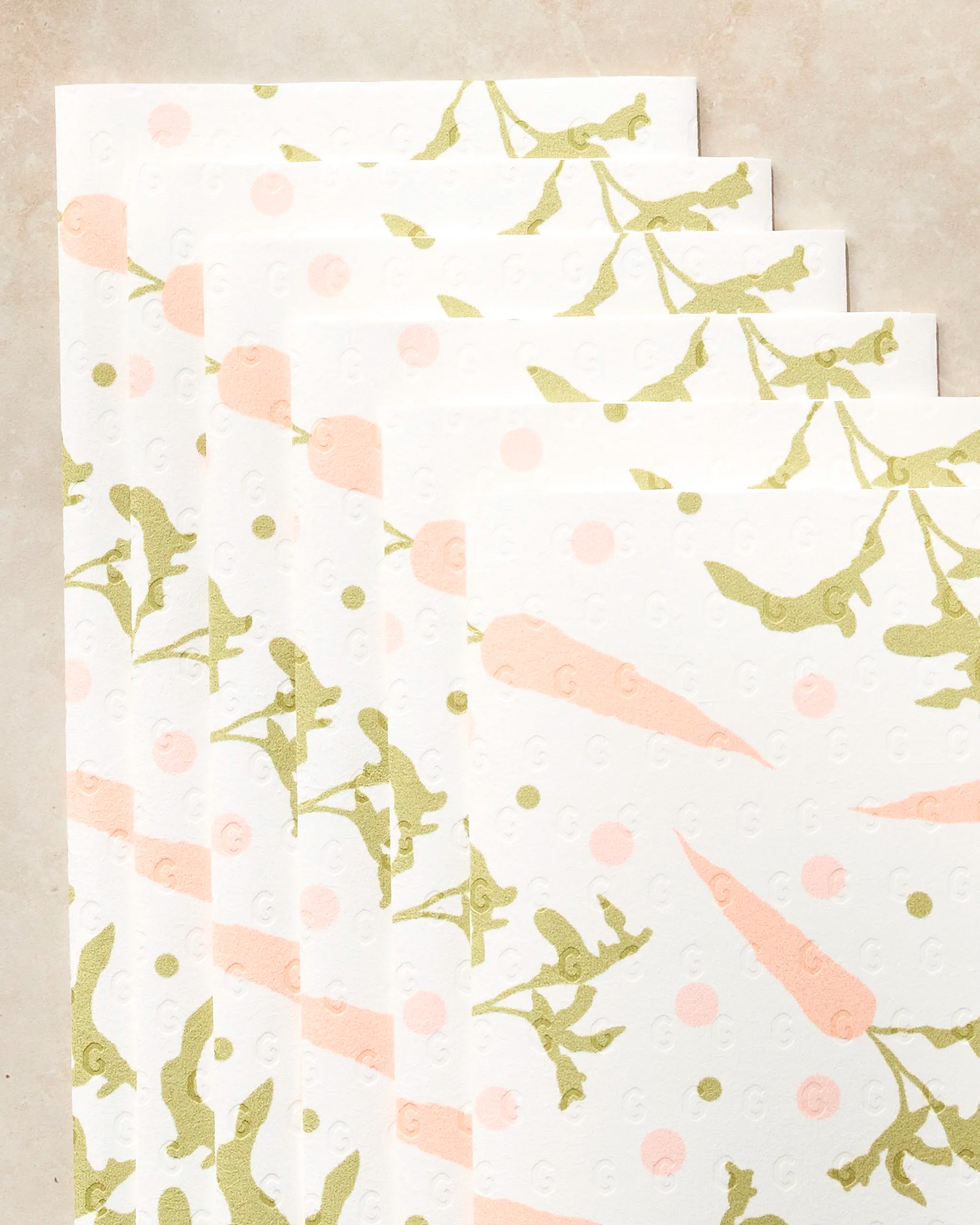 Five eco-friendly Geometry kitchen towels from the Carrot Toss collection, featuring green leafy branches and peach-colored carrots, are displayed fanned out on a beige surface.