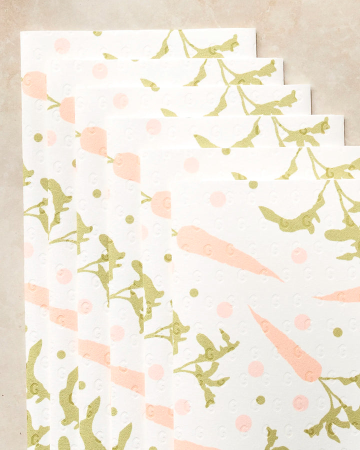 Five eco-friendly Geometry kitchen towels from the Carrot Toss collection, featuring green leafy branches and peach-colored carrots, are displayed fanned out on a beige surface.
