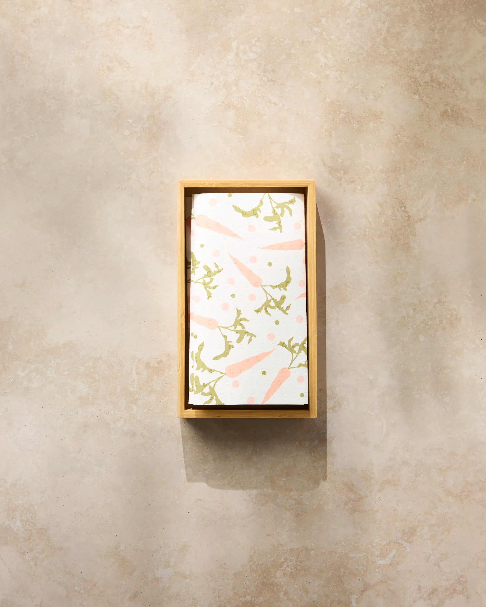 The Geometry Carrot Toss features a folded reusable paper towel with pink carrots and green leaves, displayed in a rectangular wooden tray on a beige stone surface.
