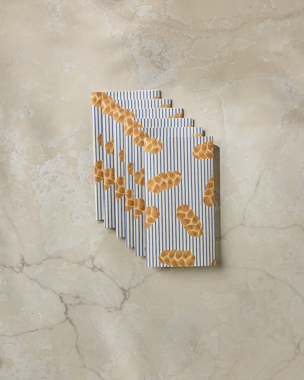 Eight “Challah Days” paper napkins from Geometry, each featuring a challah bread design, are arranged in a fanned-out stack on a light marble surface—an eco-friendly alternative to traditional serviettes that’s perfect for anyone seeking reusable paper towels for their table settings.
