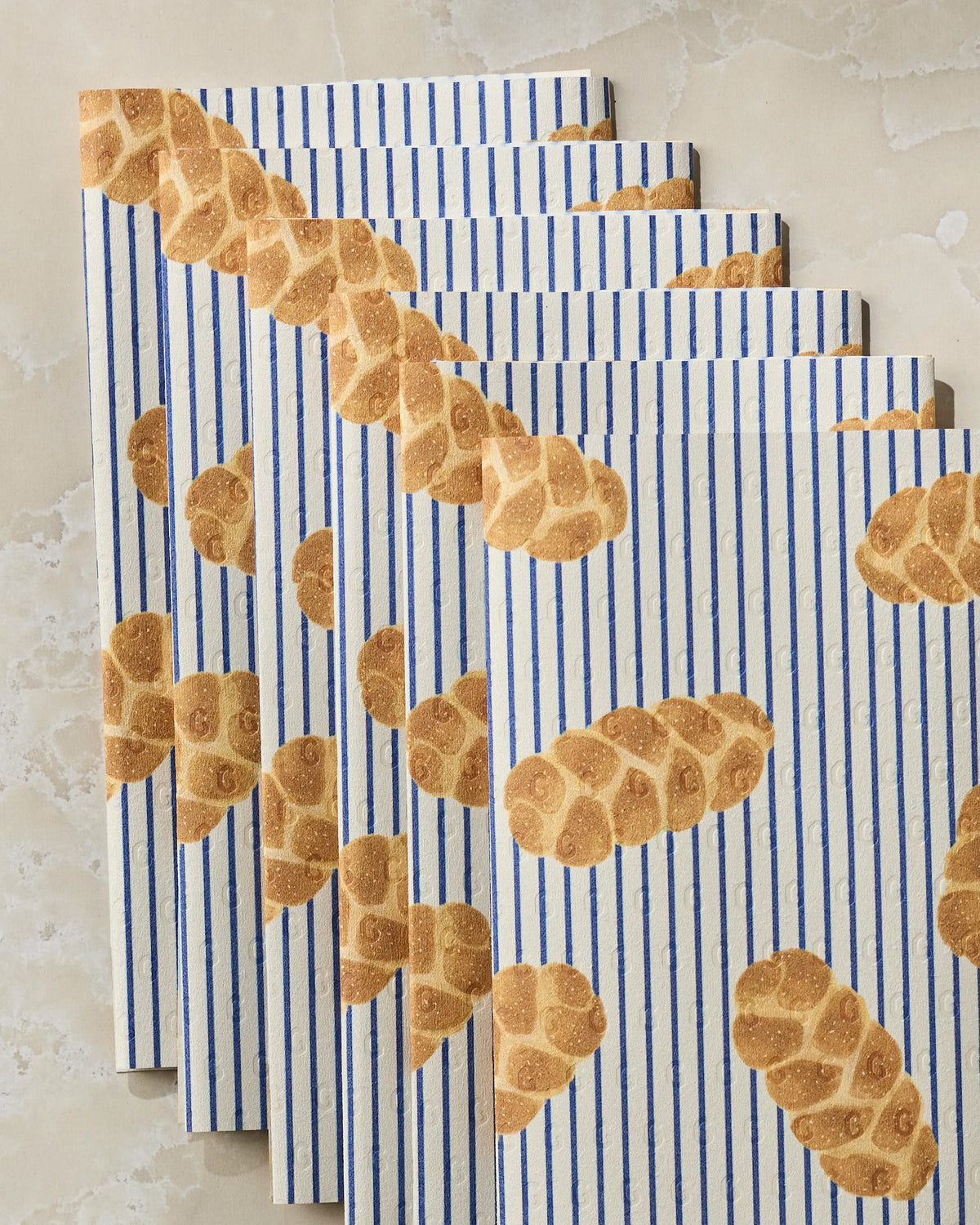 Five “Challah Days” paper bags by Geometry, featuring blue vertical stripes and braided bread illustrations, are fanned out on a light marble surface—evoking the eco-friendly style of kitchen towel packaging.