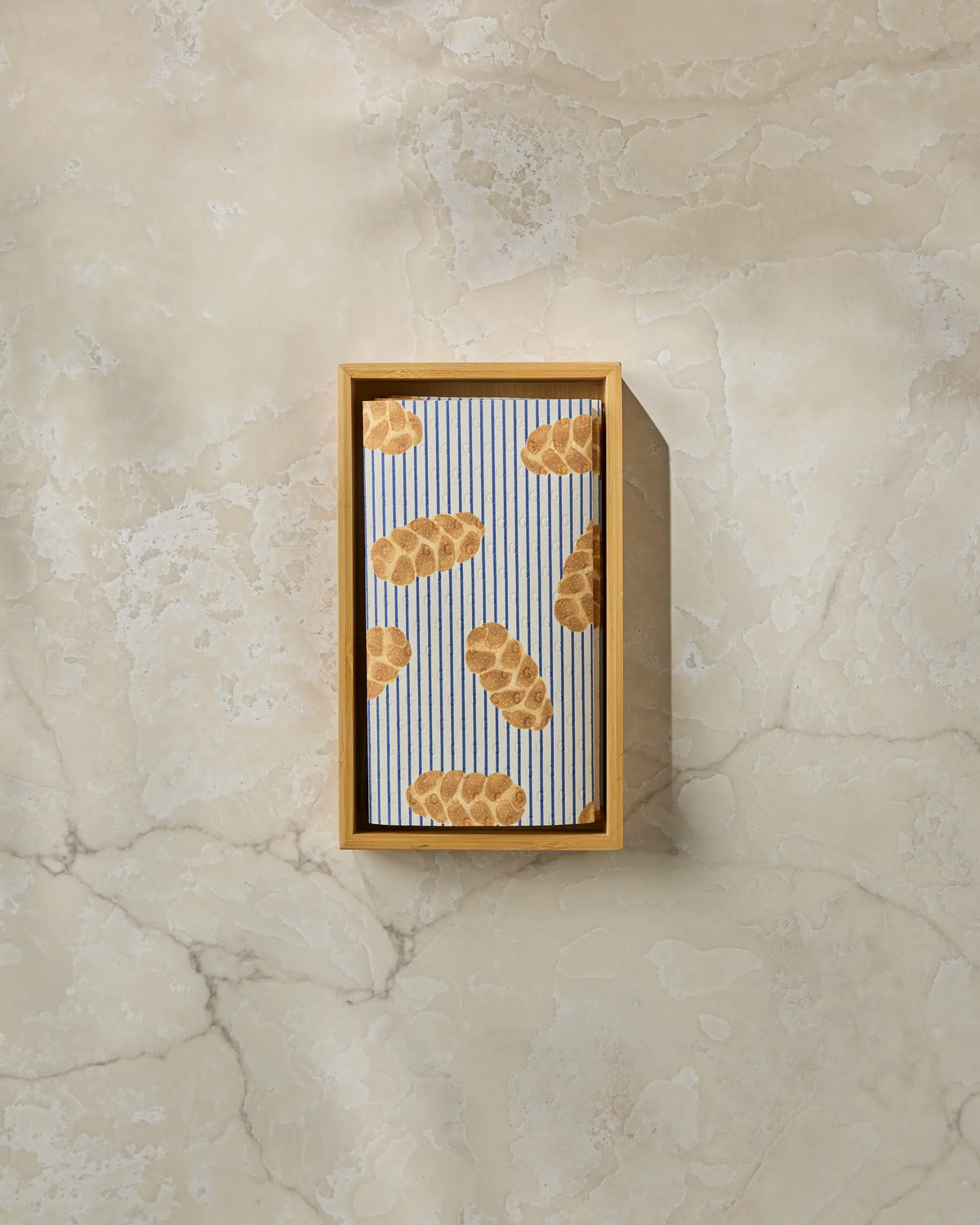 A rectangular wooden tray holds a folded Geometry “Challah Days” cloth featuring a blue and white striped pattern with brown challah loaf illustrations, resembling stylish reusable paper towels, set on a light marble surface.