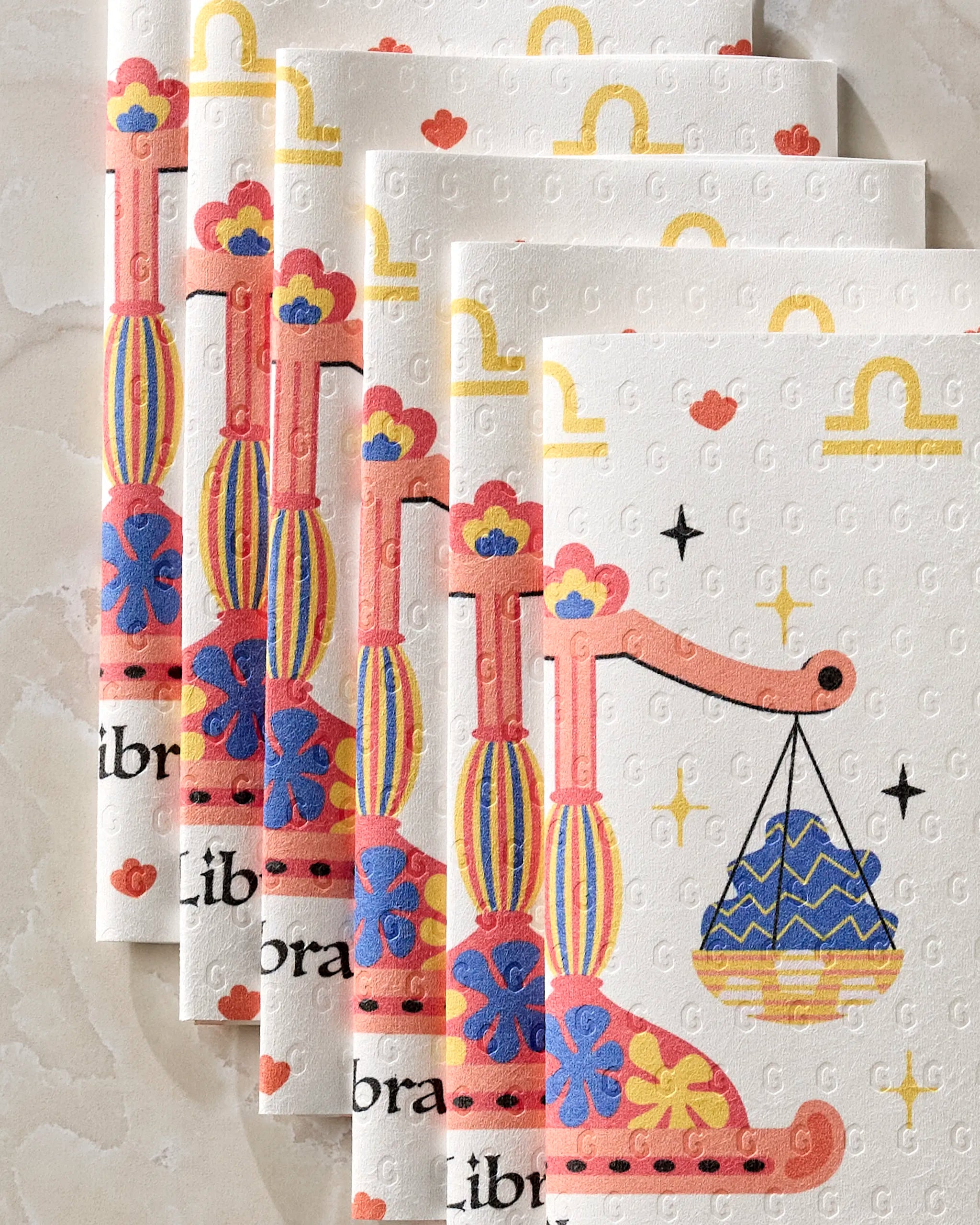 Stacked are five "Charming Libra" napkins by Heather Zhou, each with vibrant orange, blue, and yellow Libra scales on white—ideal as reusable paper towels for an eco-friendly flair.