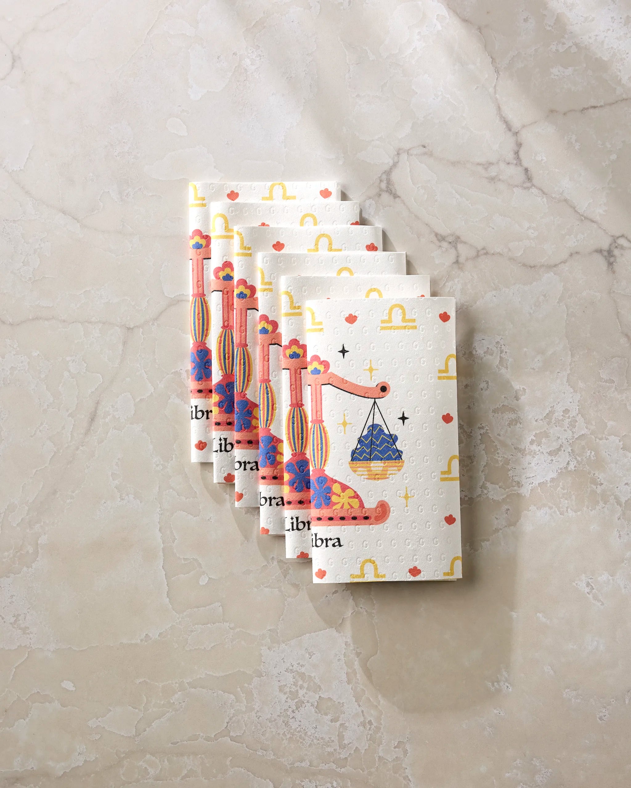 Five Charming Libra napkins by Heather Zhou, featuring a colorful scale and "libra" design, are fanned out on marble—perfect reusable paper towels for your eco-friendly kitchen.