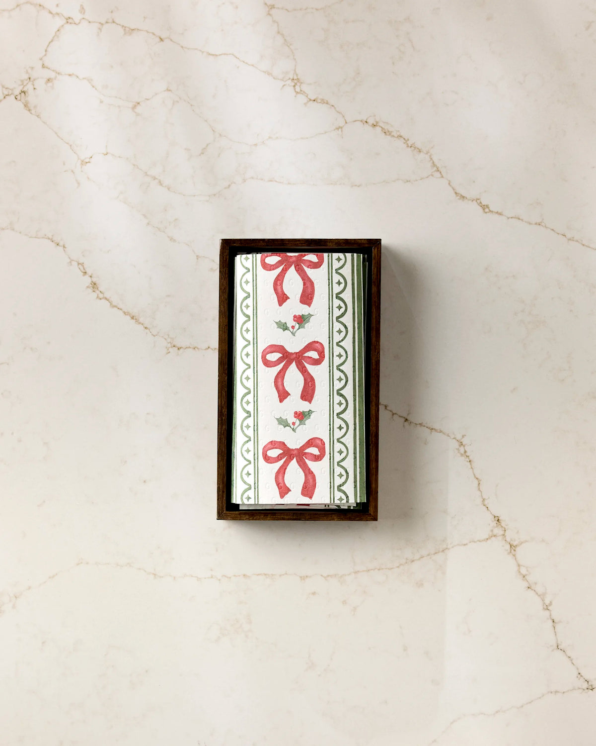 Aubrey Fairchild’s Christmas Bow Stripe: a rectangular box of eco-friendly paper towels with a white lid, red bows, green zigzag borders, and holly leaves, shown on light beige-veined marble.