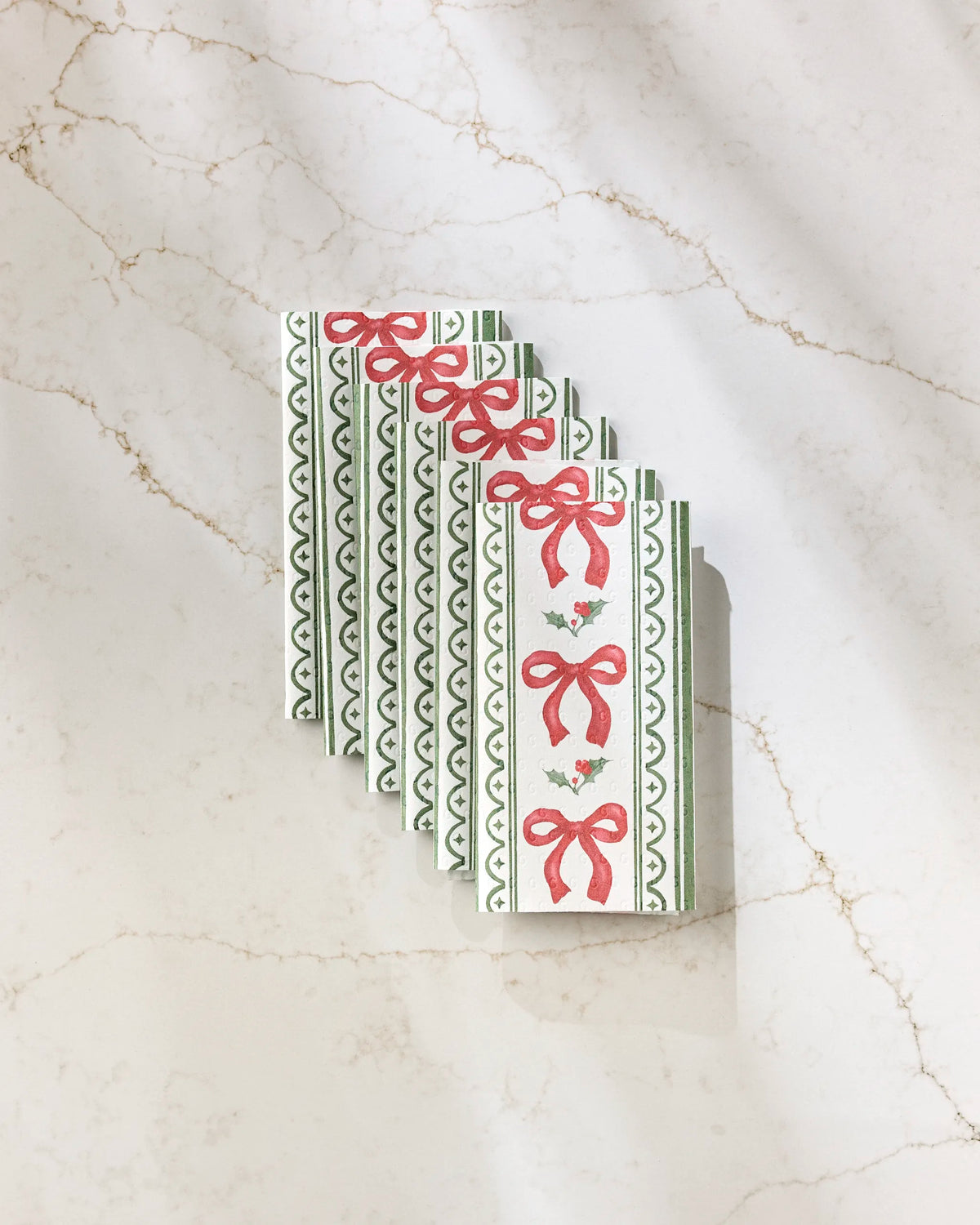 Six rectangular Aubrey Fairchild "Christmas Bow Stripe" cards, featuring green stripes and red ribbon bows, are neatly arranged in a row on a white marble surface with light brown veins. Diagonal shadows cross the image.
