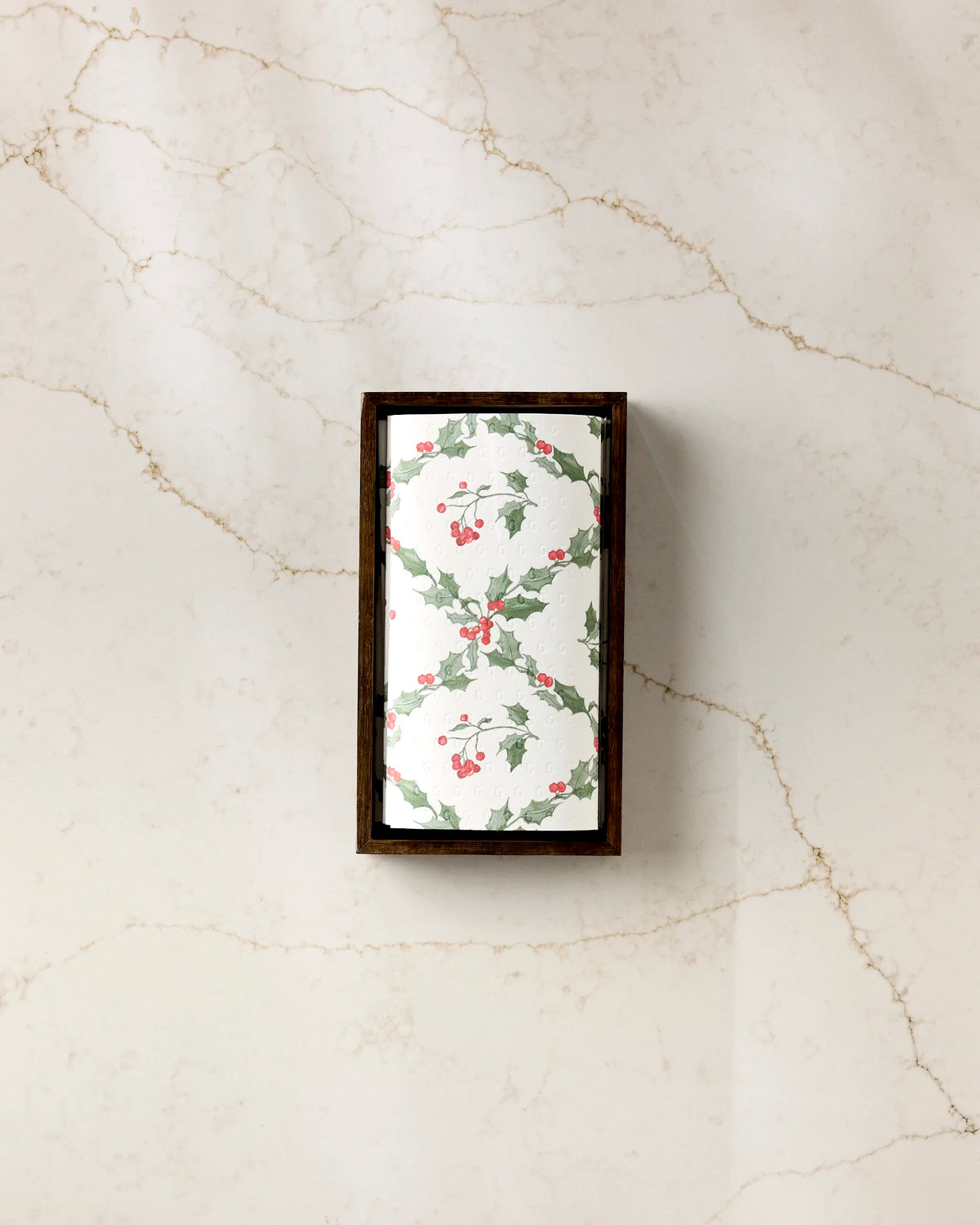 The Christmas Lattice soap by Aubrey Fairchild, wrapped in a holly and berry design, rests in a wooden tray on cream marble beside a stack of eco-friendly paper towels.