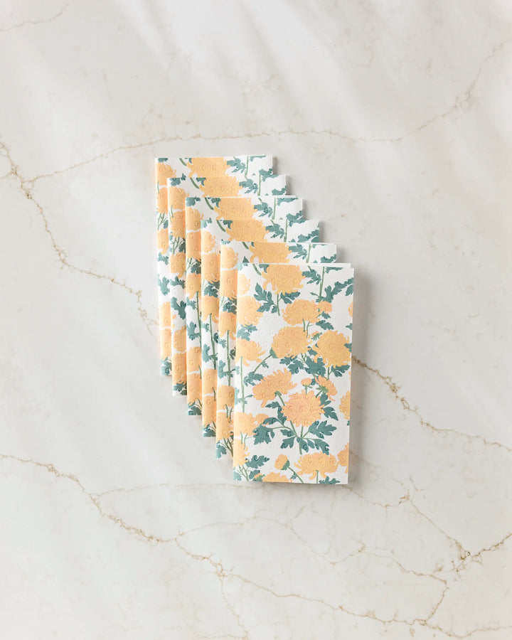 Six eco-friendly Chrysanthemum napkins by Vaishali Design Studio, featuring a yellow floral pattern with green leaves, are neatly arranged on a white marble surface with brown veining.
