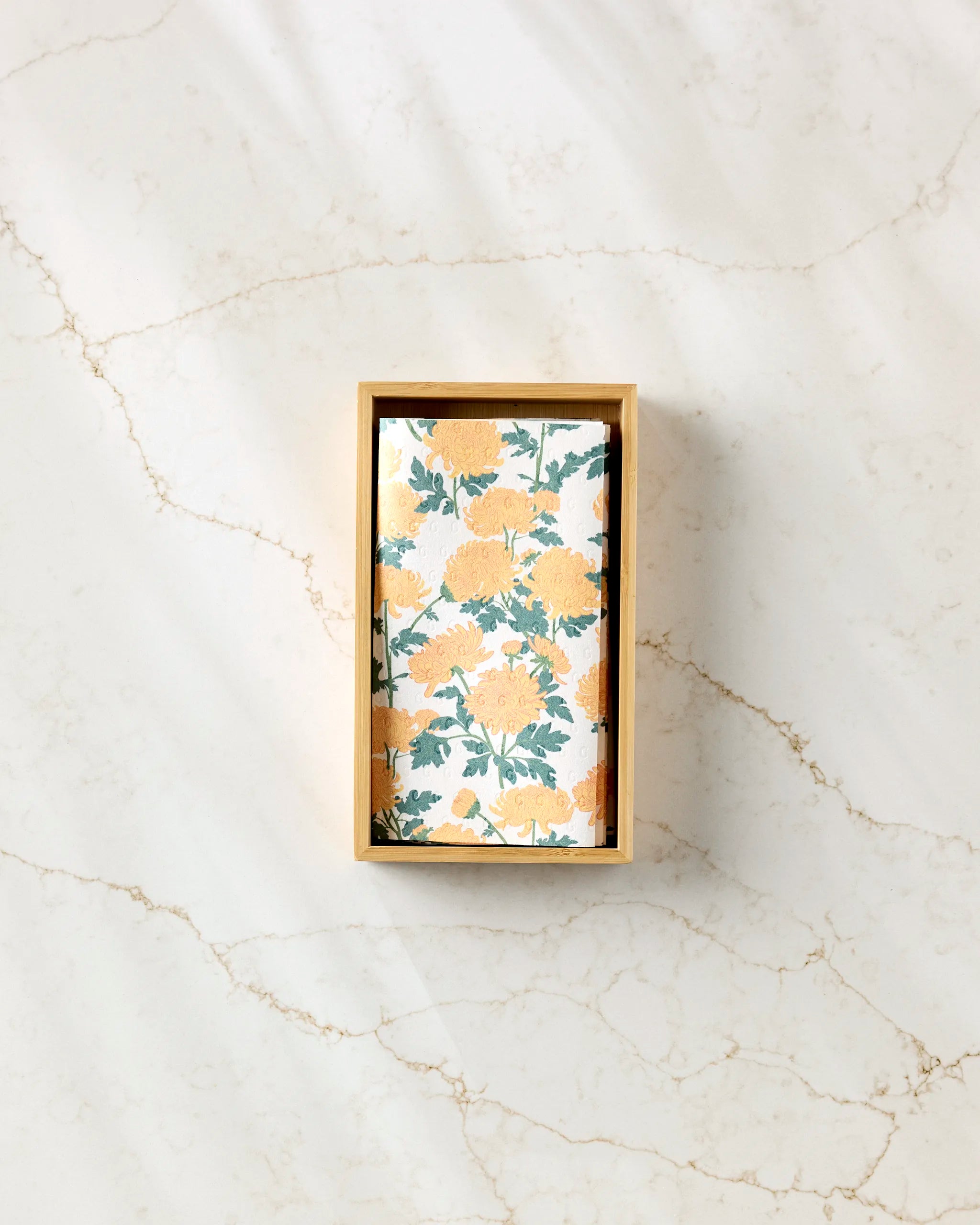 A rectangular box of eco-friendly Chrysanthemum kitchen towels by Vaishali Design Studio, featuring a yellow floral and green leaf pattern, is placed on a light marble surface with subtle brown veining.