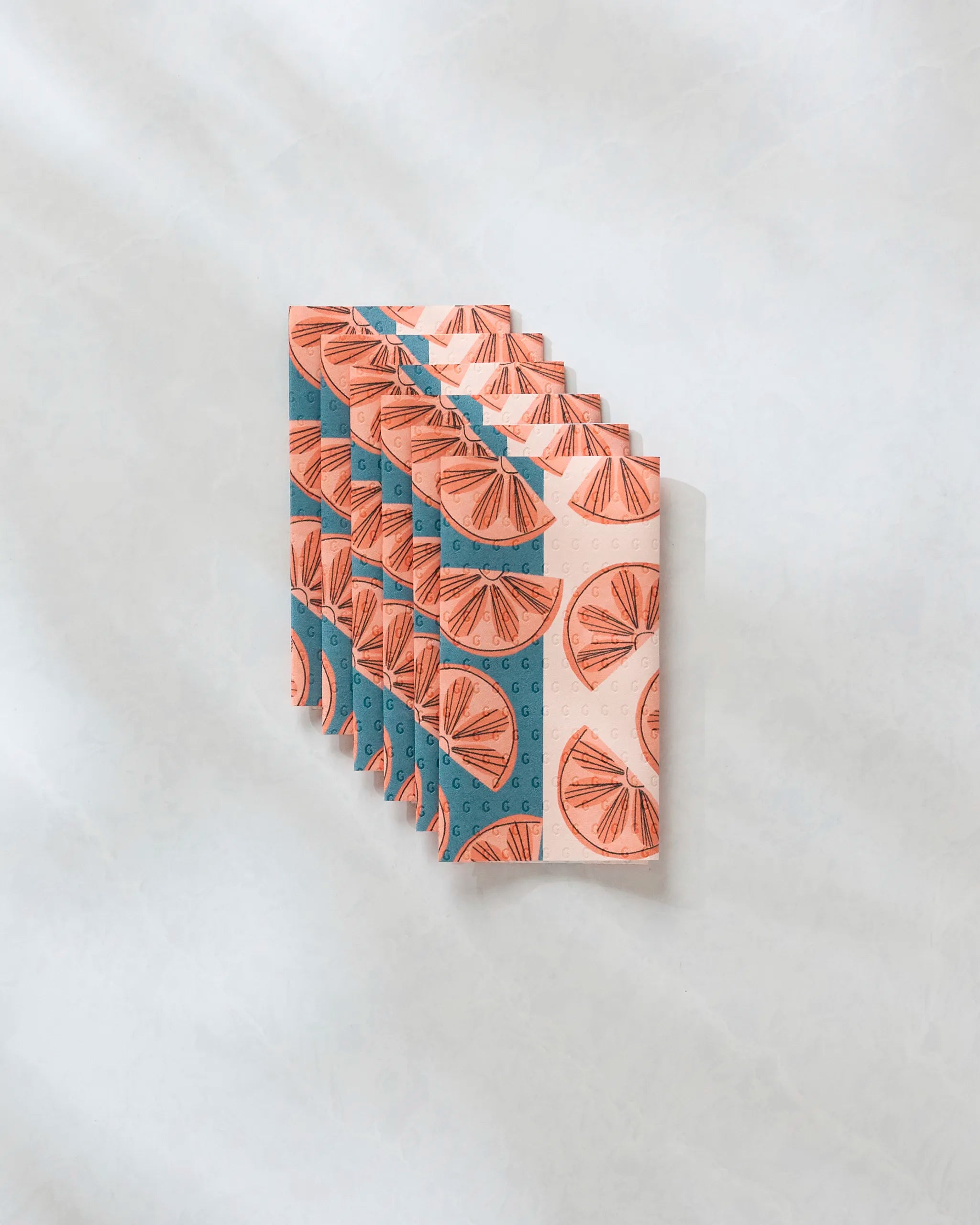 Five eco-friendly Louise Pretzel Citrus Slices kitchen towels, featuring orange slice graphics on pink and teal, are folded in a neat row on a light textured surface.