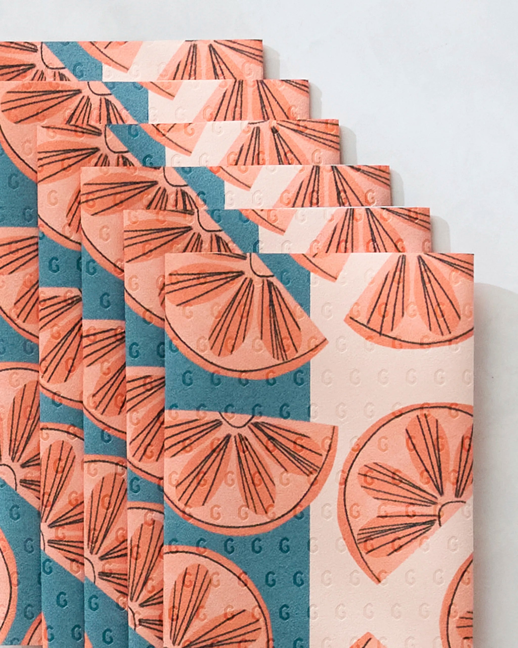 Five Louise Pretzel "Citrus Slices" notebooks, featuring orange slice illustrations and blue diagonal stripes, are fanned on a light surface. Made from post-consumer recycled materials, they offer an eco-friendly note-taking choice.