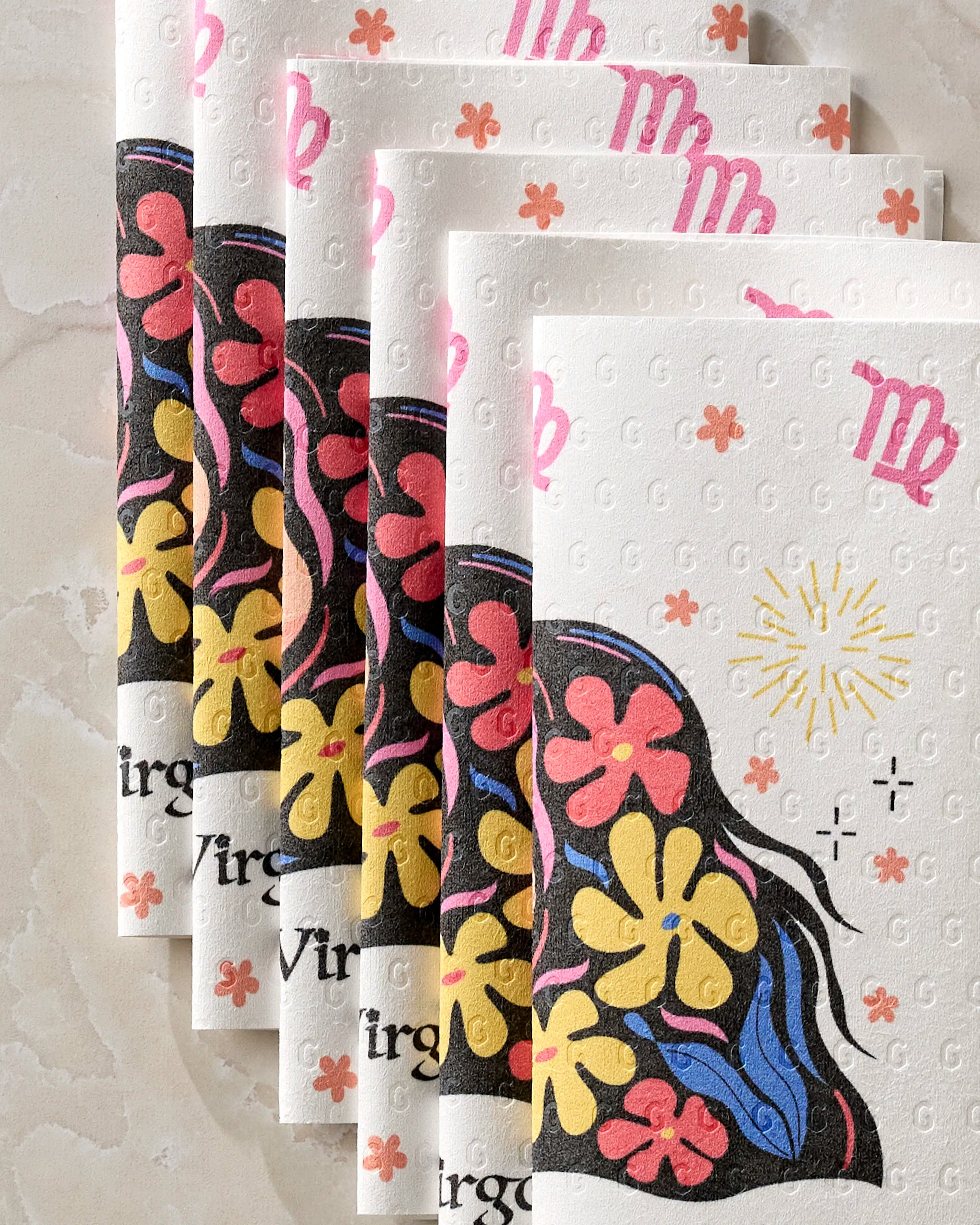 Five Clever Virgo reusable paper towels by Heather Zhou, with pink Virgo symbols and colorful flowers, are stacked on a light stone surface. These eco-friendly kitchen towels feature yellow, red, and blue floral designs.