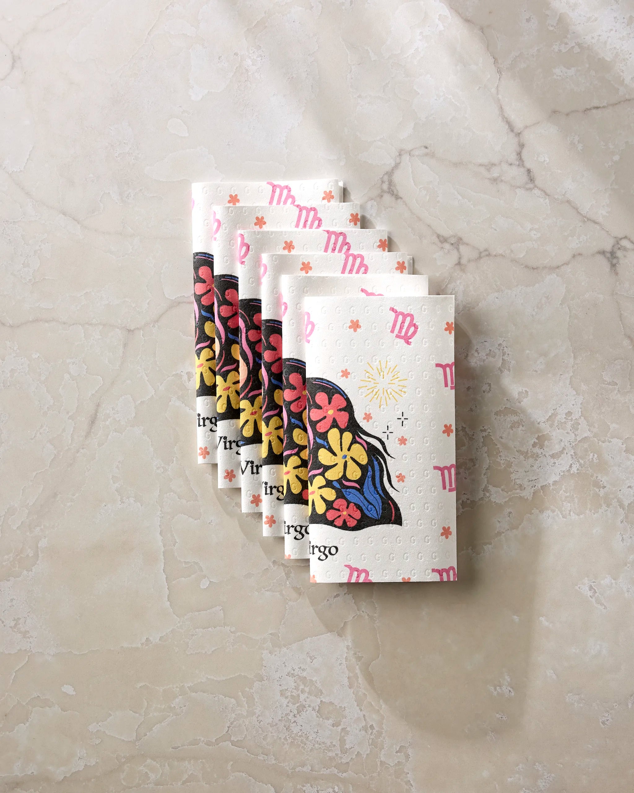 Four folded eco-friendly kitchen towels from Heather Zhou’s Clever Virgo collection, featuring a woman’s face with flowers and vibrant patterns, are arranged in a fan shape on a light marble surface.