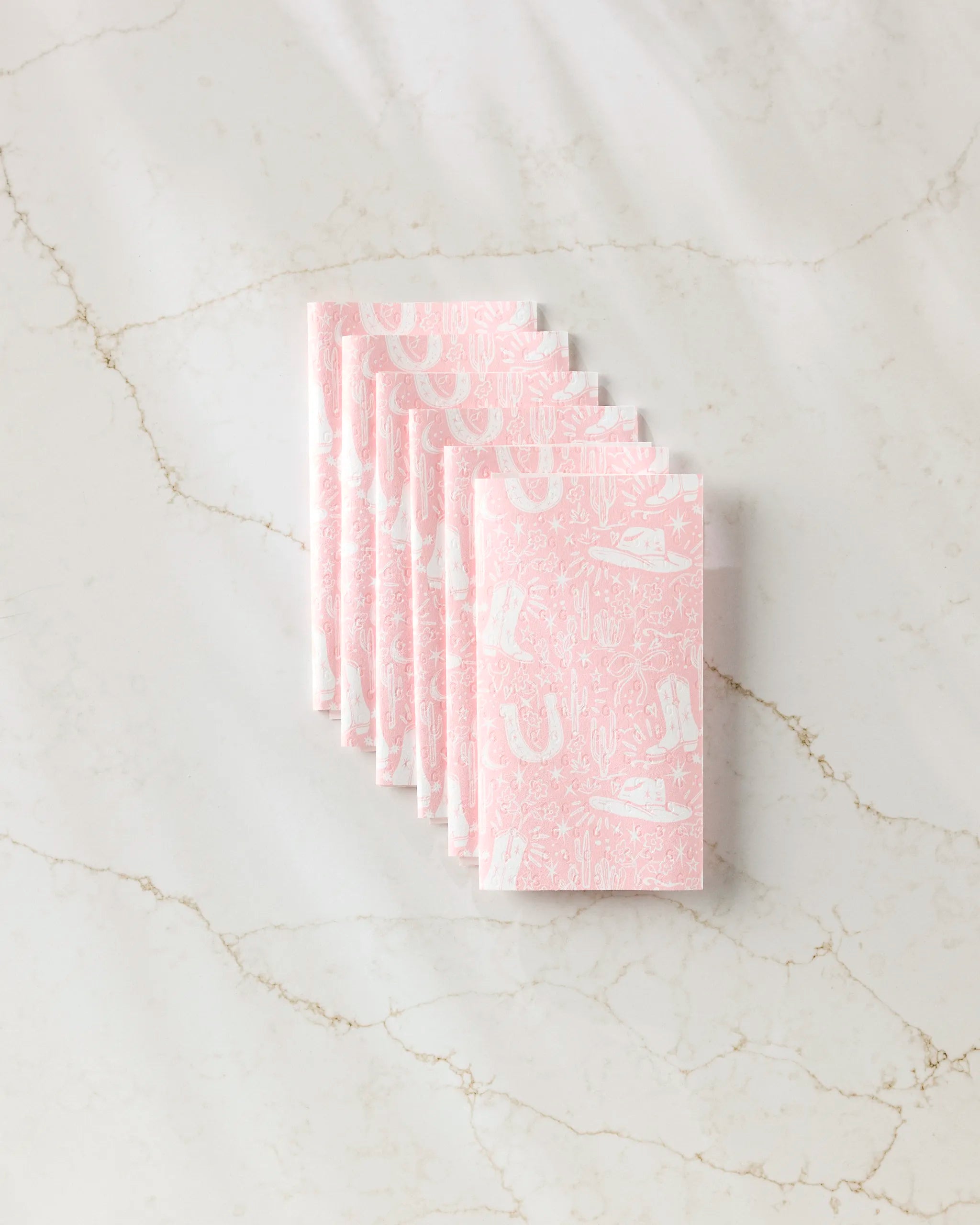 Six Cowgirl Chic reusable paper towels by Aubrey Fairchild are neatly stacked on a white marble surface, featuring pink and white western-themed patterns like boots, hats, cacti, and horseshoes.