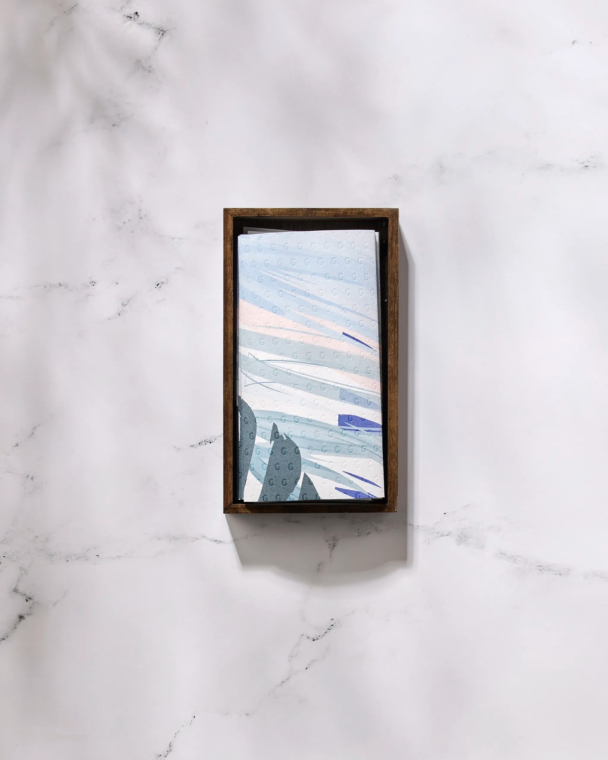 A rectangular wooden box on a white marble surface holds GEOMETRY’s Create eco-friendly kitchen towels—Not Paper Towels—featuring a light blue patterned cloth with abstract shapes, perfect for sustainable home routines.