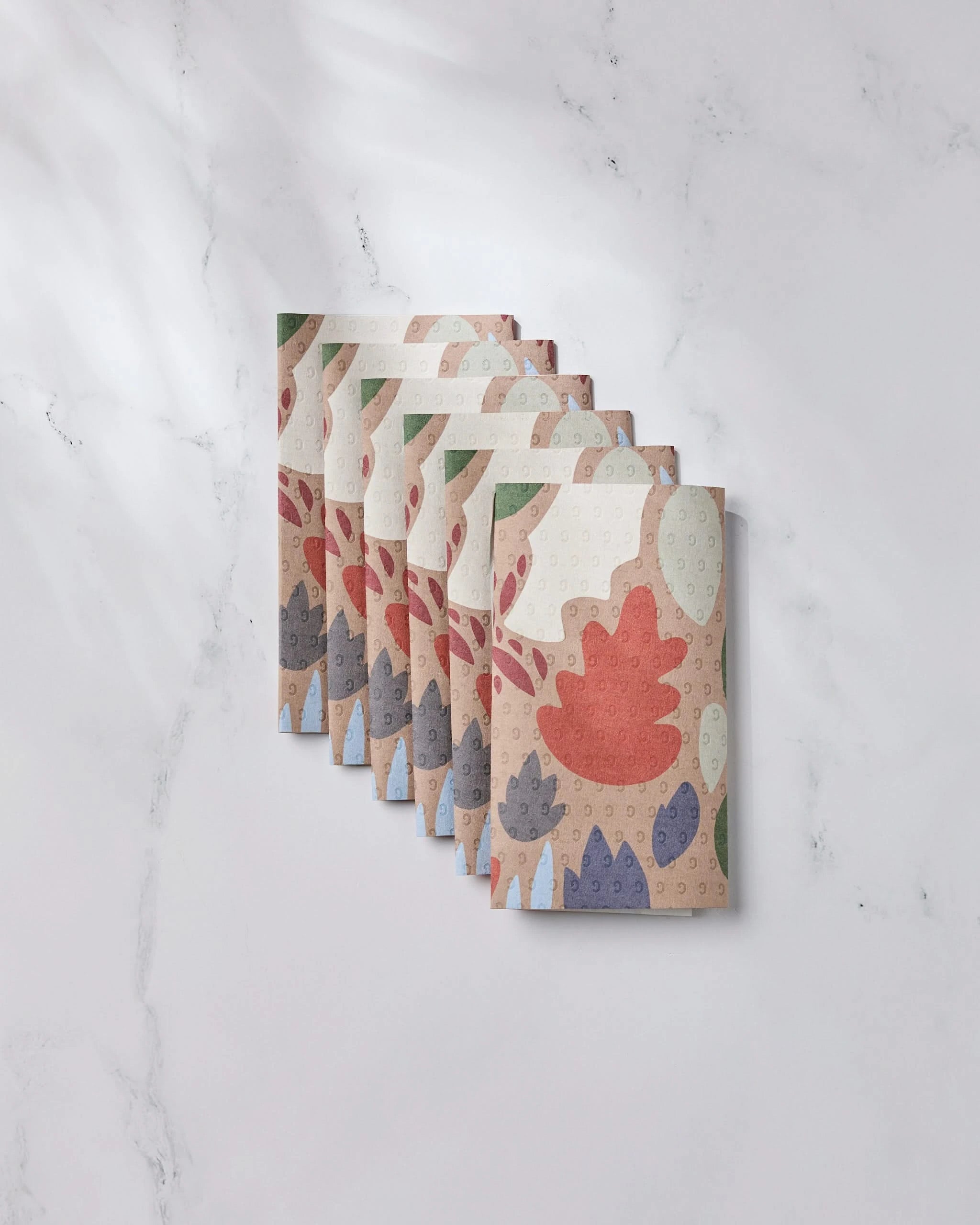 Six rolls of Geometry’s Cut Paper Leaves Mocha toilet paper, featuring a colorful leaf pattern and made from post-consumer recycled materials, are arranged neatly on a white marble surface.