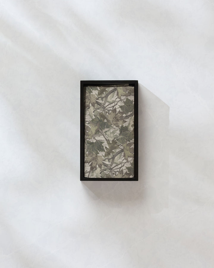 Geometry's "Dad Camo" features a rectangular black frame and a camouflage-inspired pattern of green and brown leaves, evoking reusable paper towels. It's displayed on a light, textured wall with soft shadows.