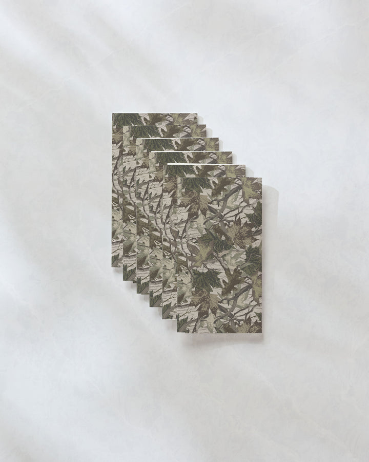A stack of six Geometry Dad Camo cards with a camouflage pattern rests on a light, textured surface resembling eco-friendly paper towels, with soft shadows cast to the right.