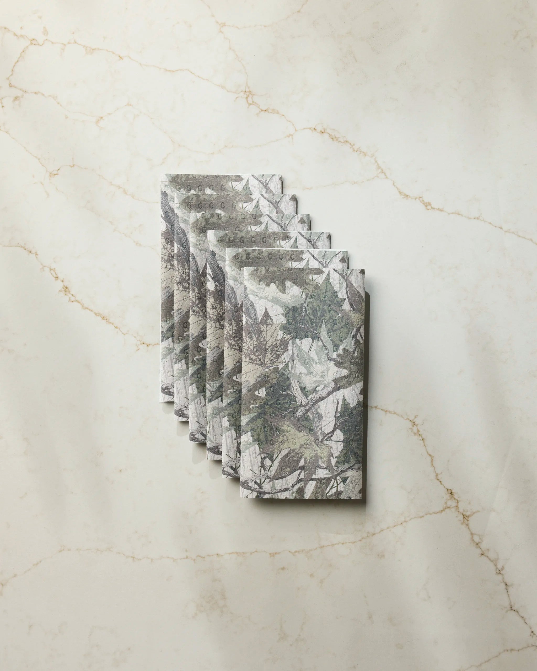 Six Geometry Dad Camo reusable paper towels, featuring gray and green patterns, are neatly stacked on a light-colored marble surface with subtle veining.