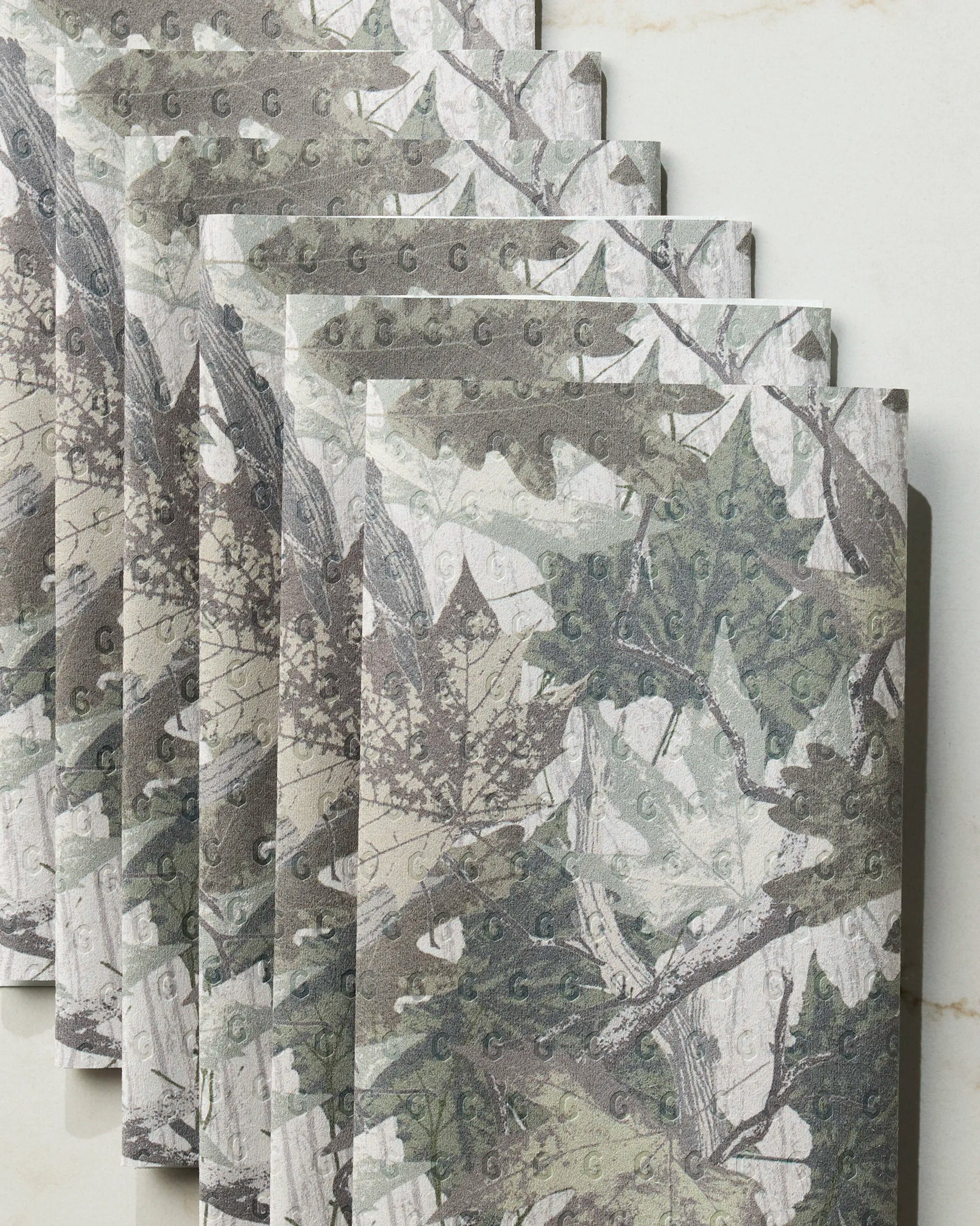 Five overlapping Geometry Dad Camo reusable paper towels, featuring a green and brown camouflage leaf and branch pattern on a light background, are displayed on a marble-like surface.