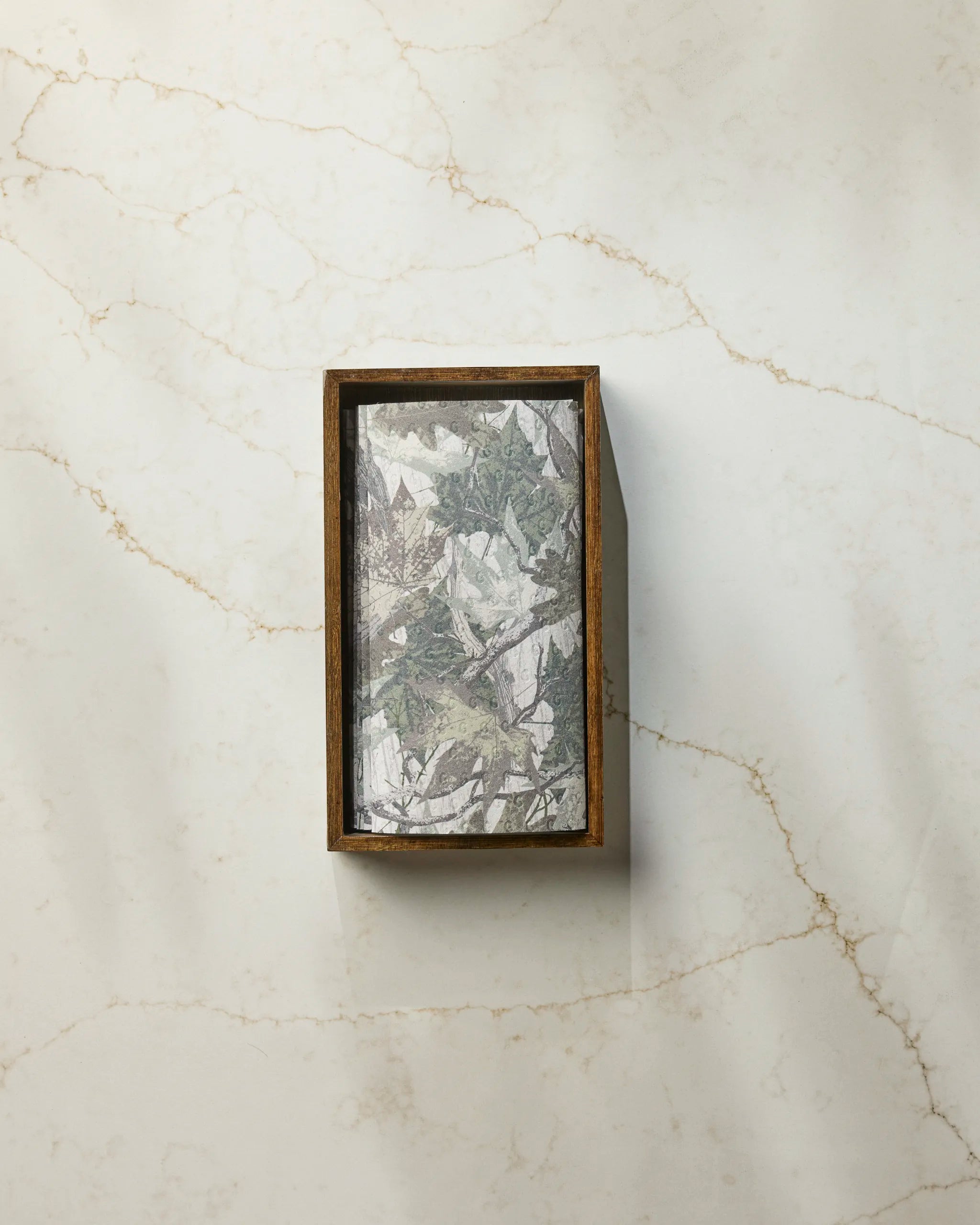 The Dad Camo by Geometry, a rectangular wooden box with leafy patterned paper like eco-friendly kitchen towels, rests on a light marble surface with subtle brown veins.