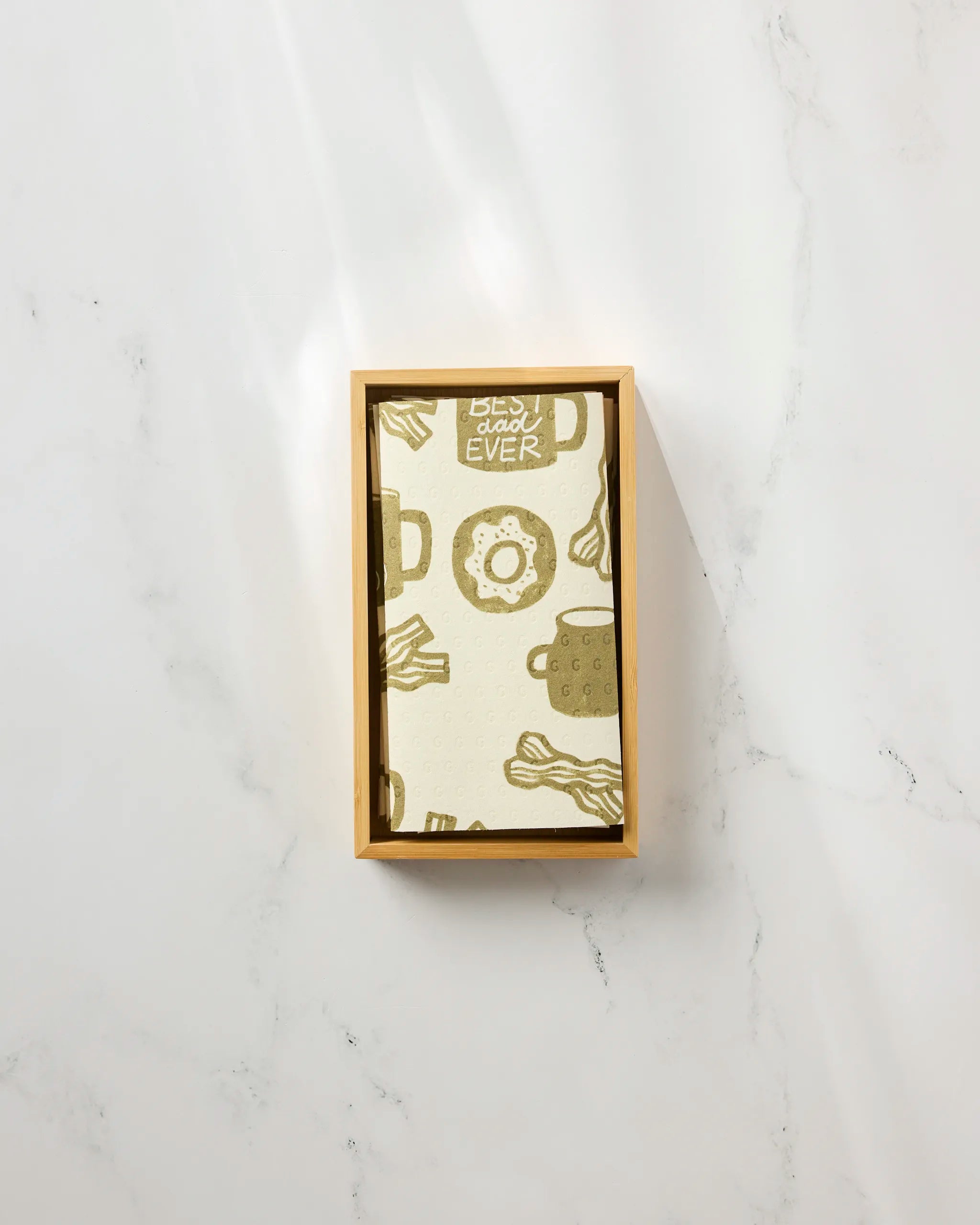 A rectangular "Dad Fuel" card by Lisa Nelson, illustrated with bacon, eggs, coffee mugs, and toast, sits in a wooden tray on white marble next to eco-friendly kitchen towels. The card reads "Best Dad Ever" at the top center.