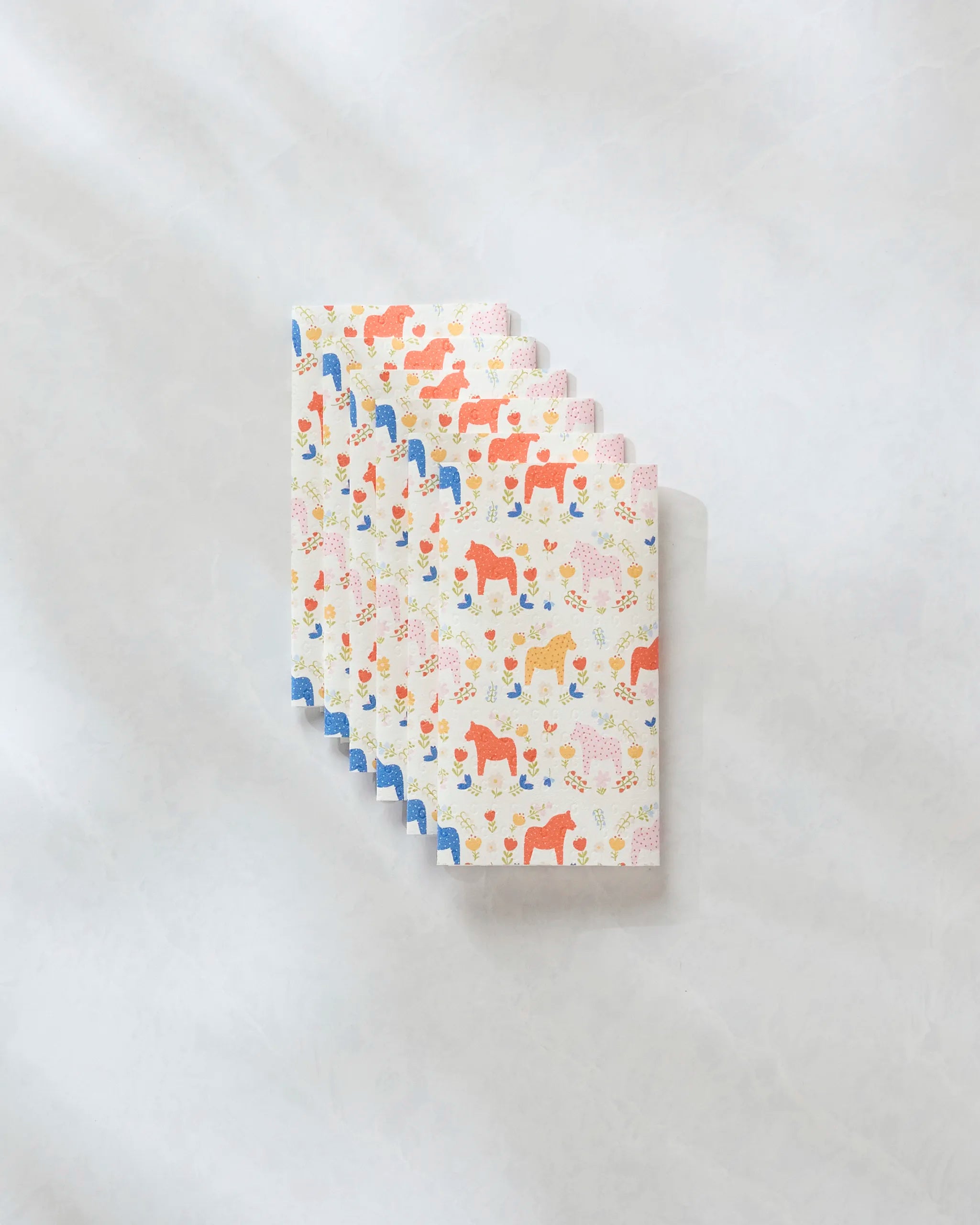 A stack of Sara Mari Dala Garden Floral eco-friendly kitchen towels, featuring red, blue, and orange horse and flower patterns on a light background, sits on a pale marble surface.
