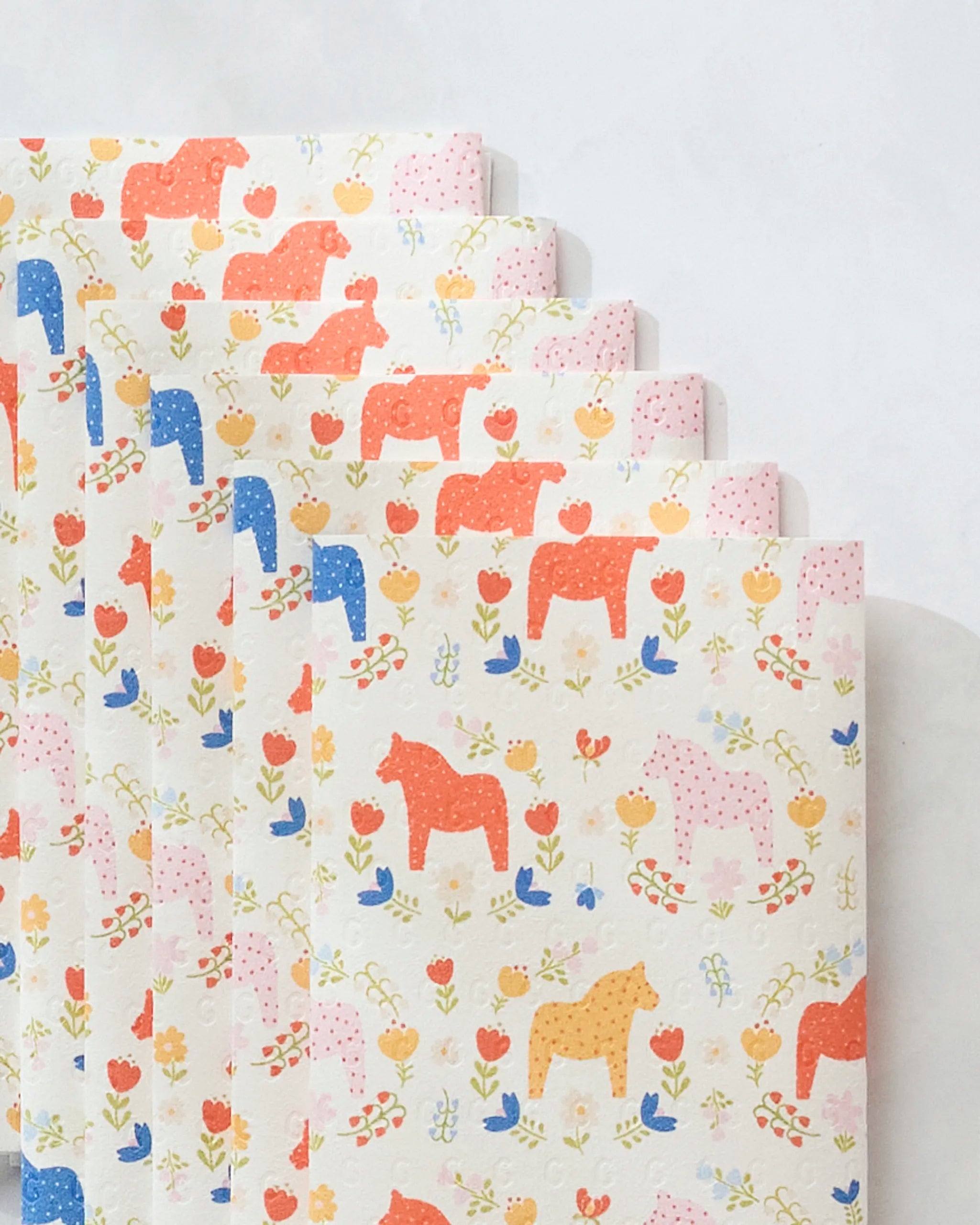 Six sheets of Sara Mari's Dala Garden Floral paper towels are fanned out, showing eco-friendly white sheets with colorful Dala horse designs in red, blue, yellow, and pink among small flowers and hearts.