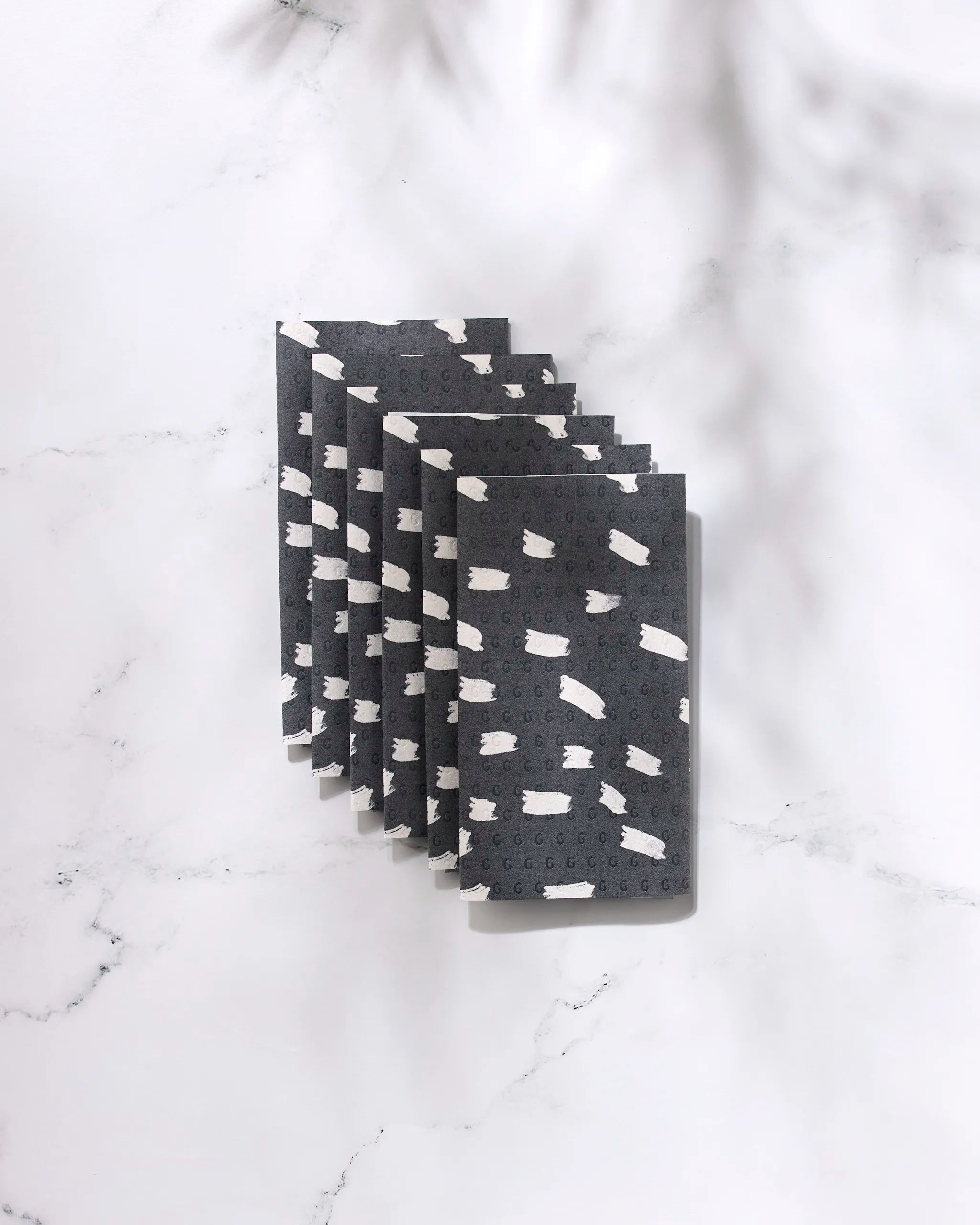Six "Dark Out" rectangular sponges by GEOMETRY, featuring black surfaces with white abstract patterns inspired by eco-friendly kitchen towels, are neatly stacked on a white marble surface, casting soft shadows.