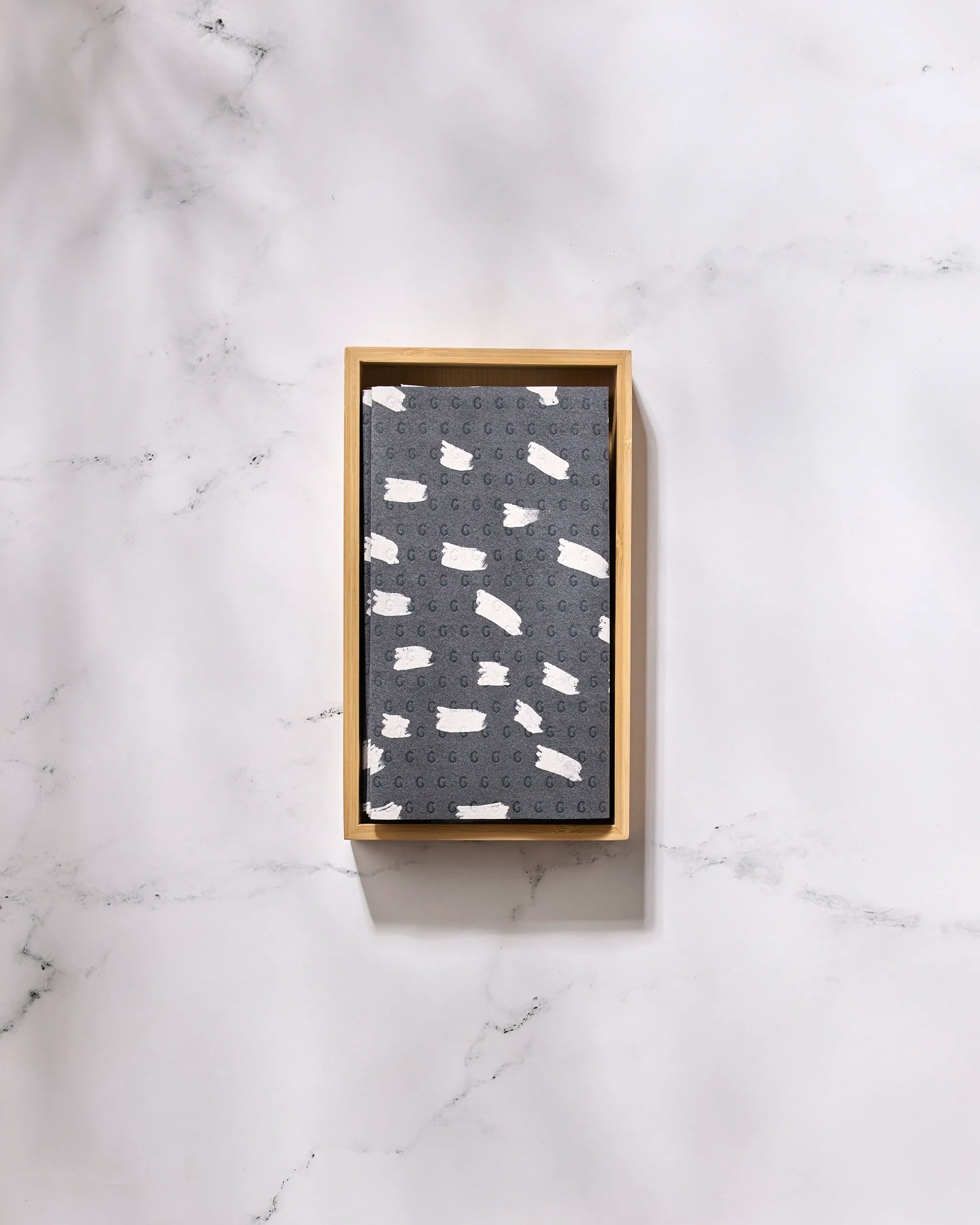 A rectangular wooden tray holds GEOMETRY's Dark Out terrazzo-style notebook with white and gray speckles, plus eco-friendly paper towels, all arranged on a white marble surface.