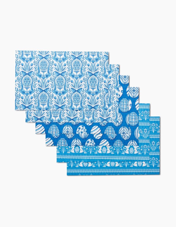 Four rectangular Delft Blue Easter Eggs and Bunnies placemats by Sarah Daugherty are made from eco-friendly paper towels, arranged in a staggered stack, and showcase intricate blue and white floral, botanical, and geometric designs.