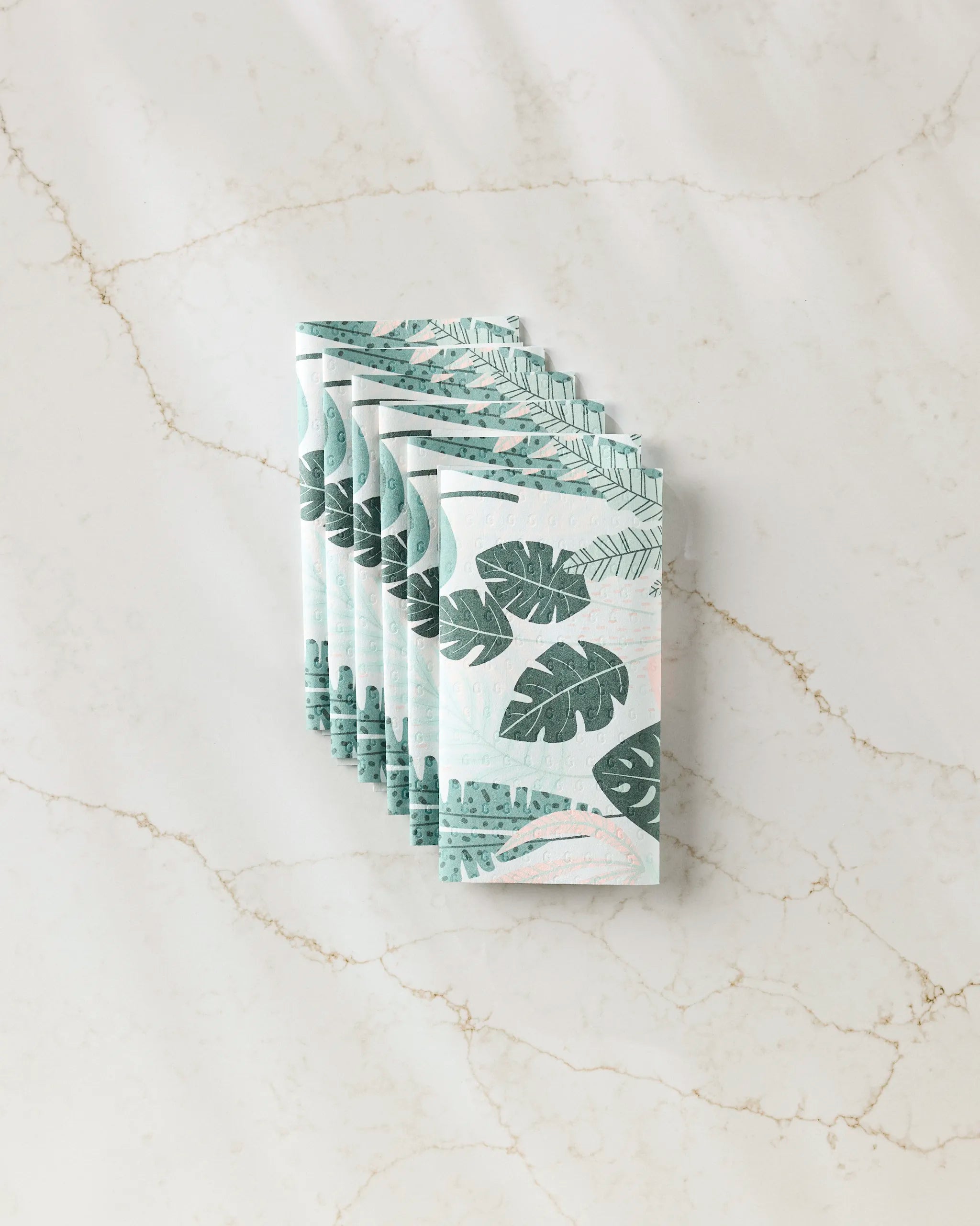 Five neatly stacked GEOMETRY Diedre recycled paper towels, featuring green tropical leaf patterns, are displayed on a light marble surface with subtle veining.
