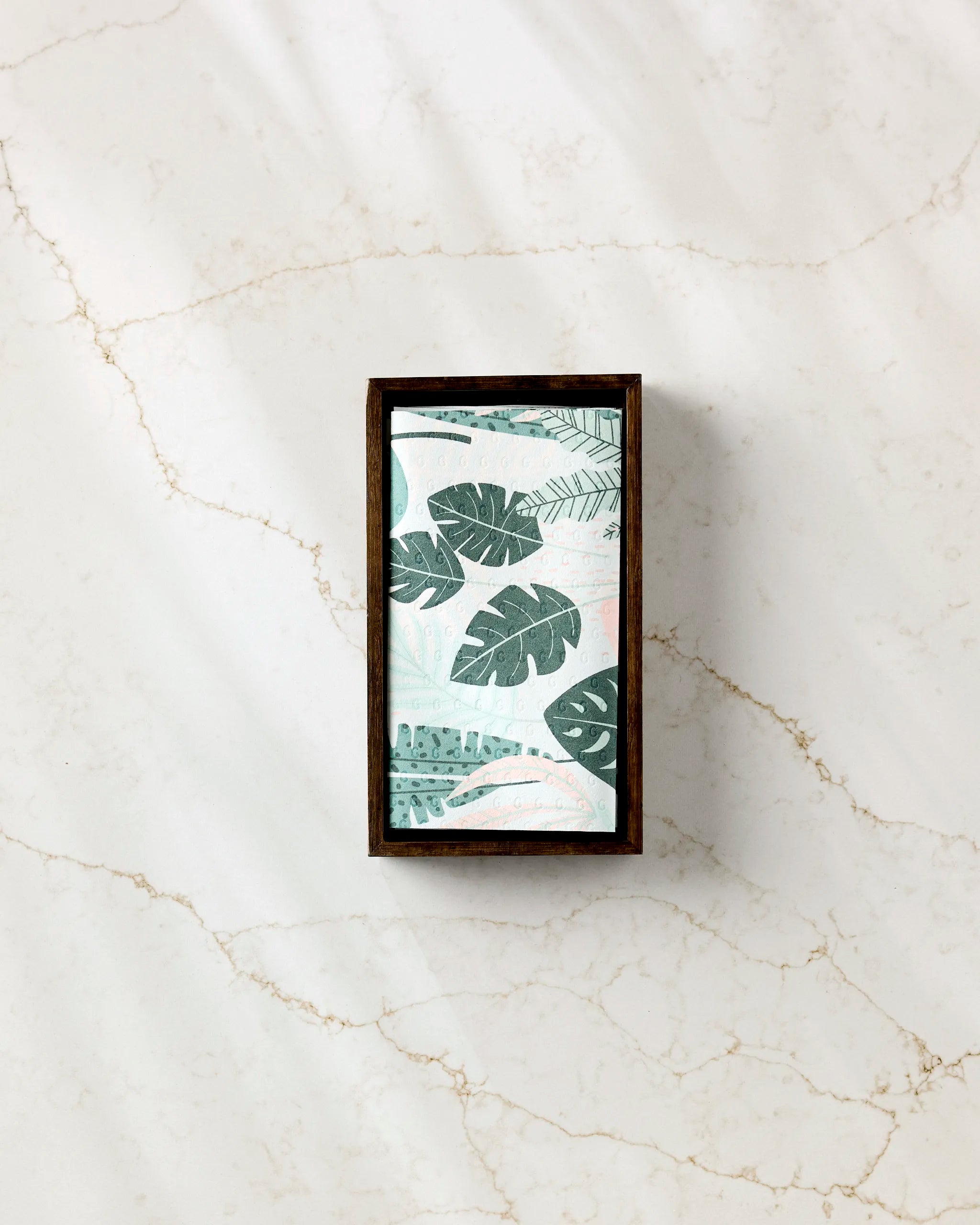 Diedre by GEOMETRY—a rectangular framed artwork featuring green tropical leaves on a light background—rests atop a white marble surface with beige veins, its soft shadows evoking the elegance of eco-friendly paper towels.